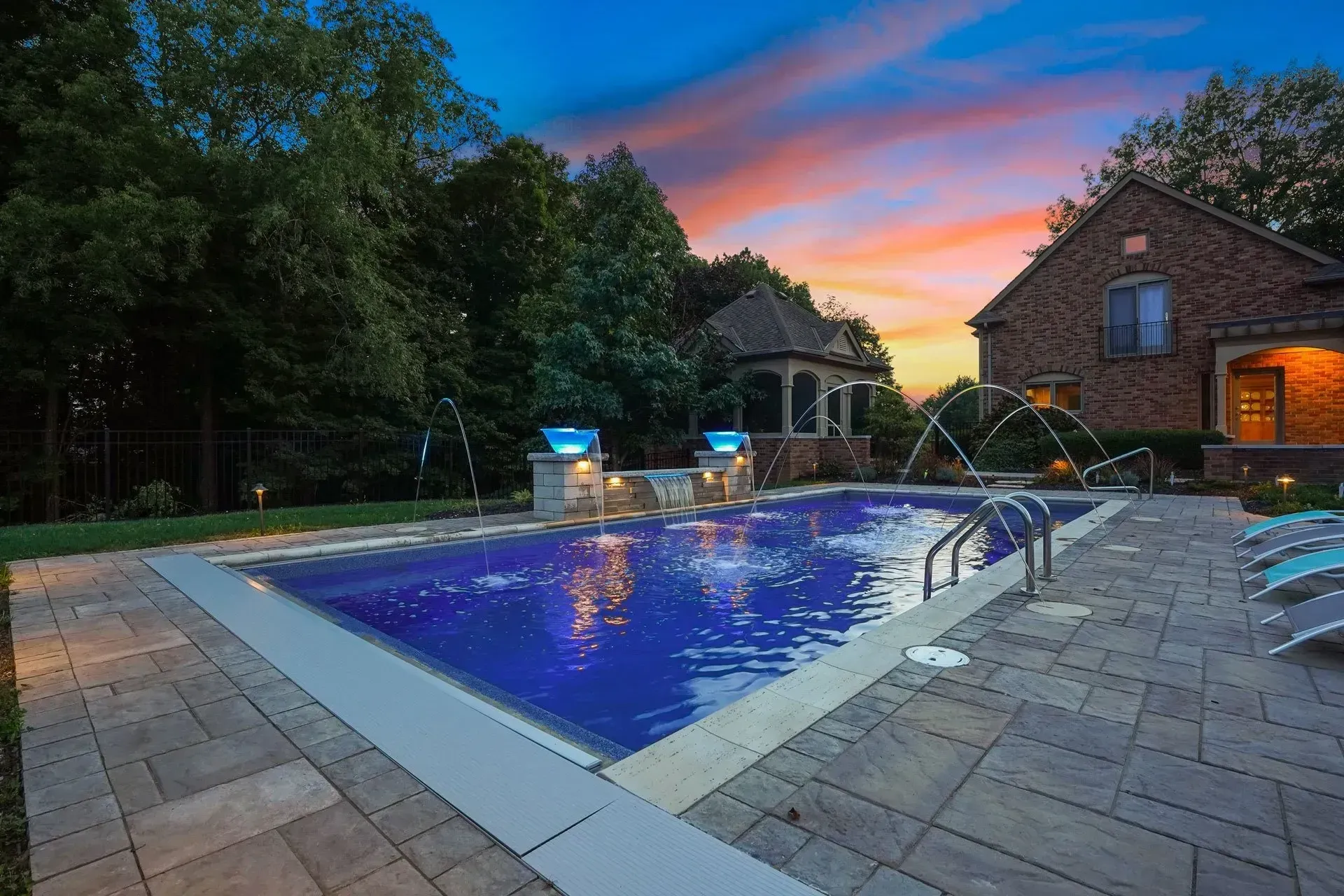 A backyard pool at dusk with water features illuminated by blue lights, set against a colorful sunset.
