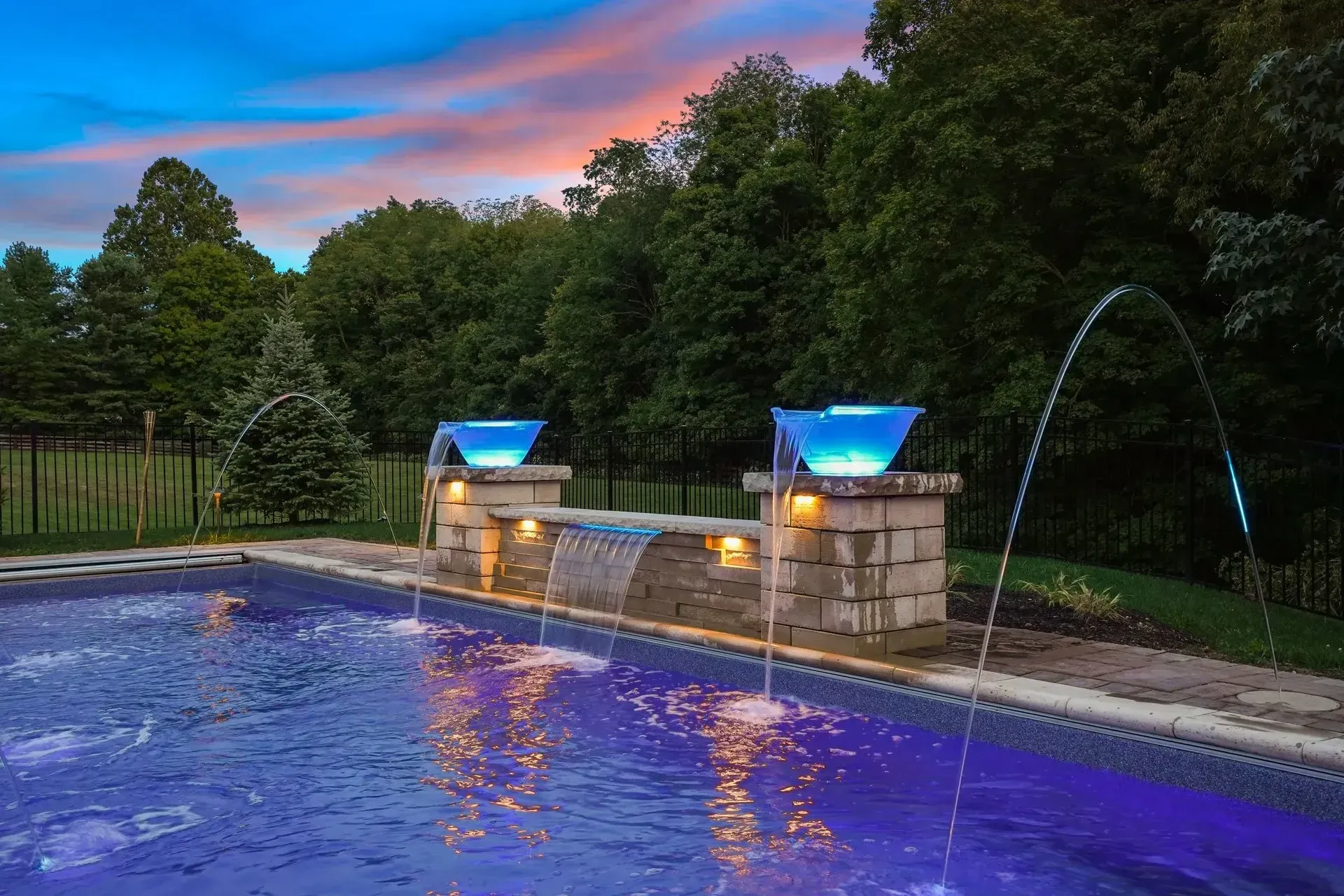 Pool with fountains, a brick water feature, and blue-lit accents at dusk. Trees and a colorful sky in the background.
