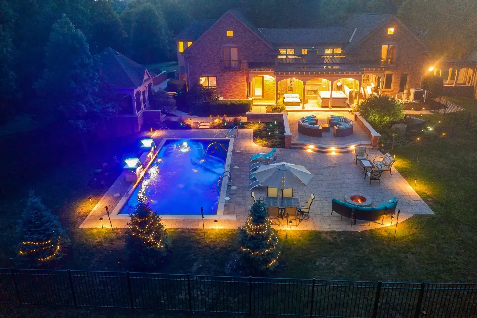 Nighttime aerial view of a luxury home with a lit-up pool, patio, and manicured lawn. The house is brick with warm interior lighting.