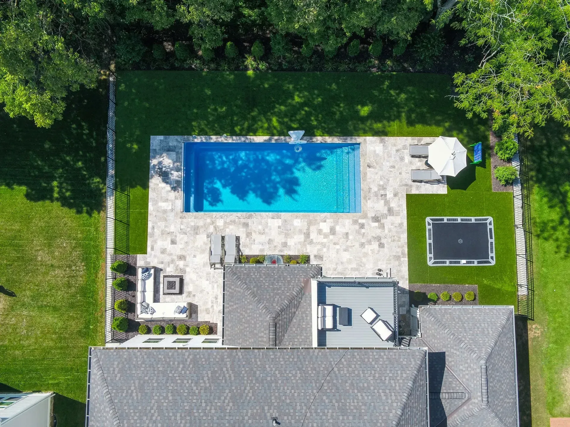 Aerial view of a backyard with a rectangular pool, patio, trampoline, and a house with a grey roof.