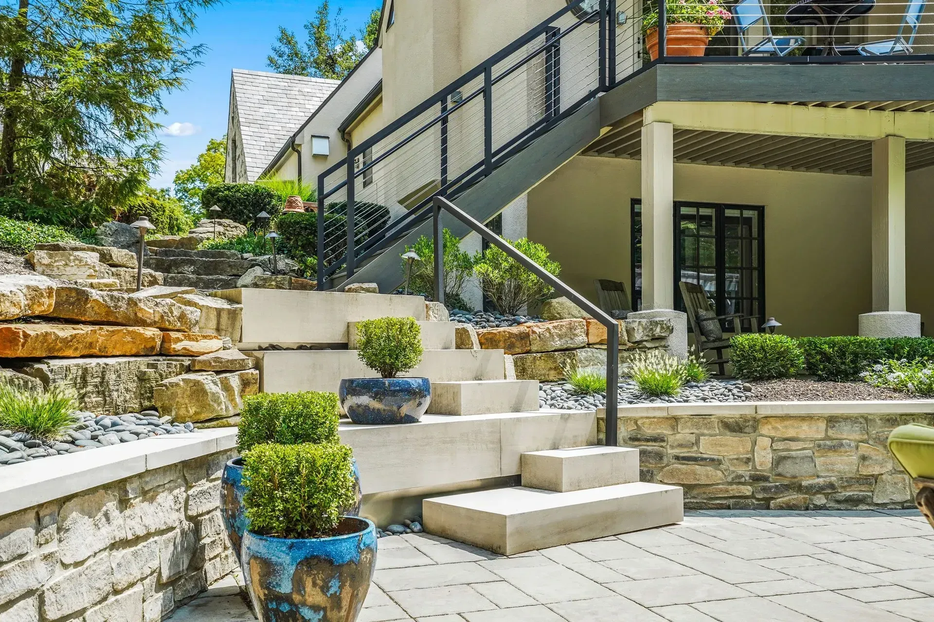 Stone staircase with modern metal railing leads to a second-story deck. The stairs are flanked by stone landscaping and decorative potted shrubs.