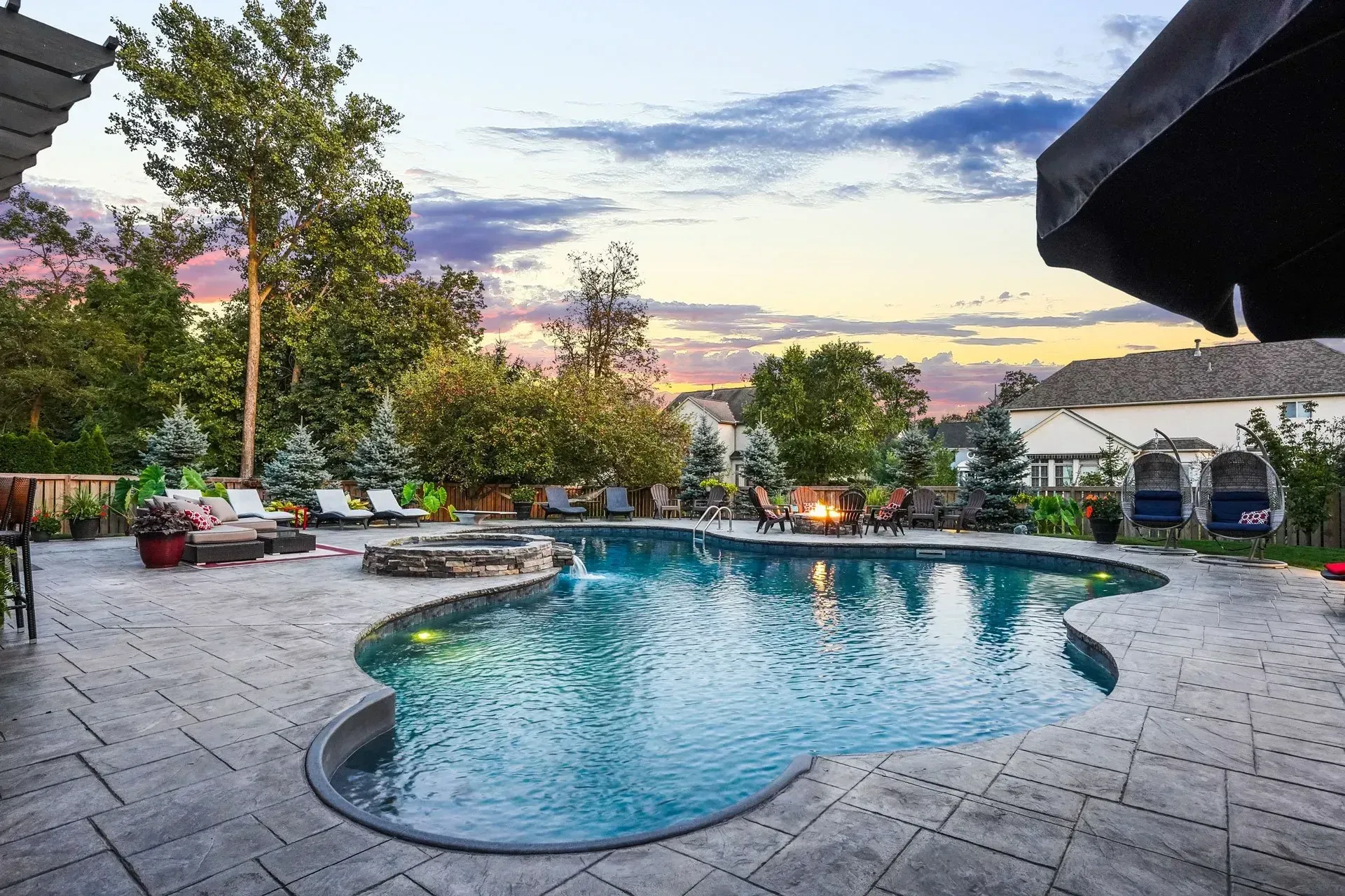 Backyard pool area with a flowing pool, patio, fire pit, and lounge seating under a colorful sky at dusk.