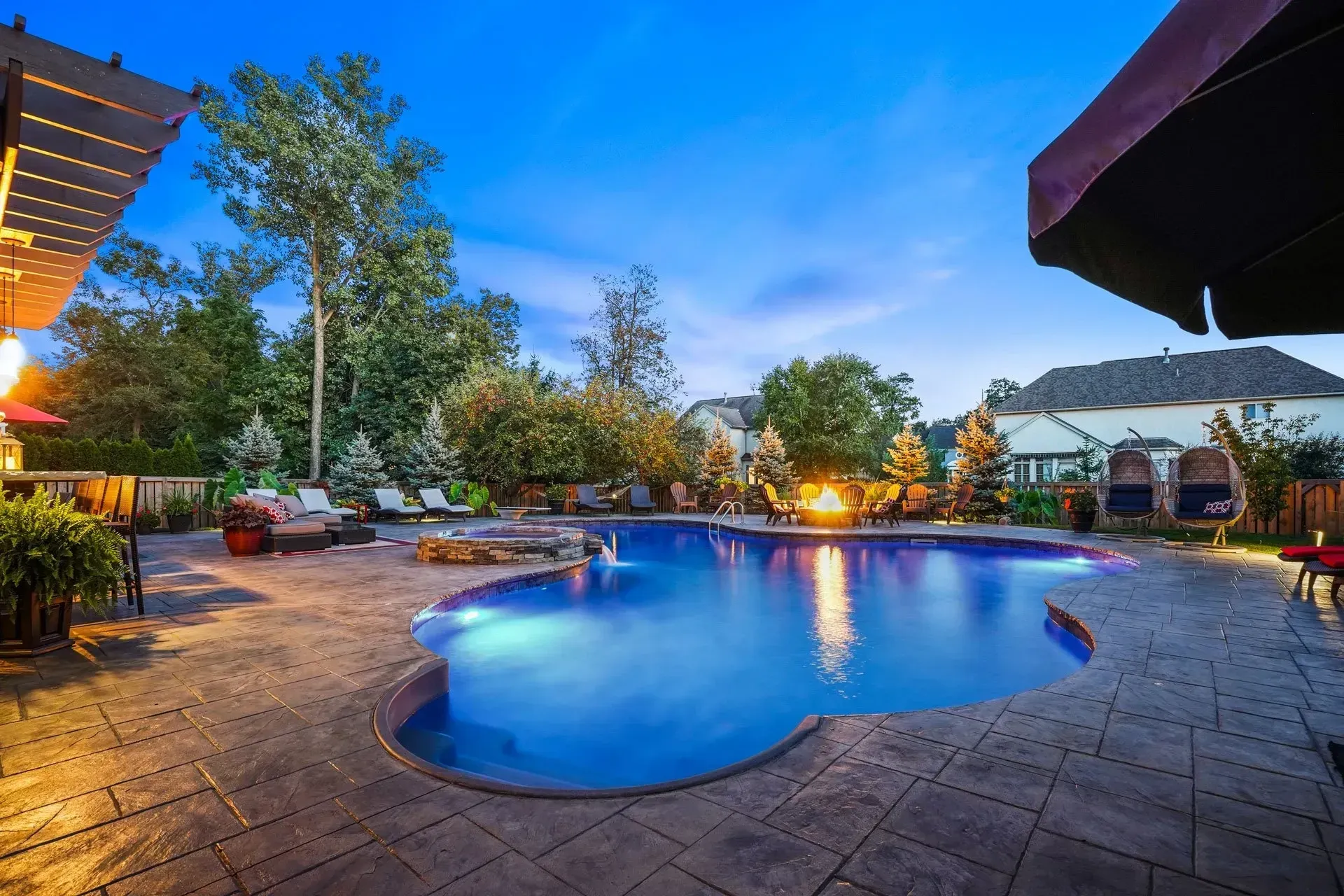 Nighttime backyard scene with a pool lit up with blue lights, surrounded by patio furniture and landscaping under a twilight sky.