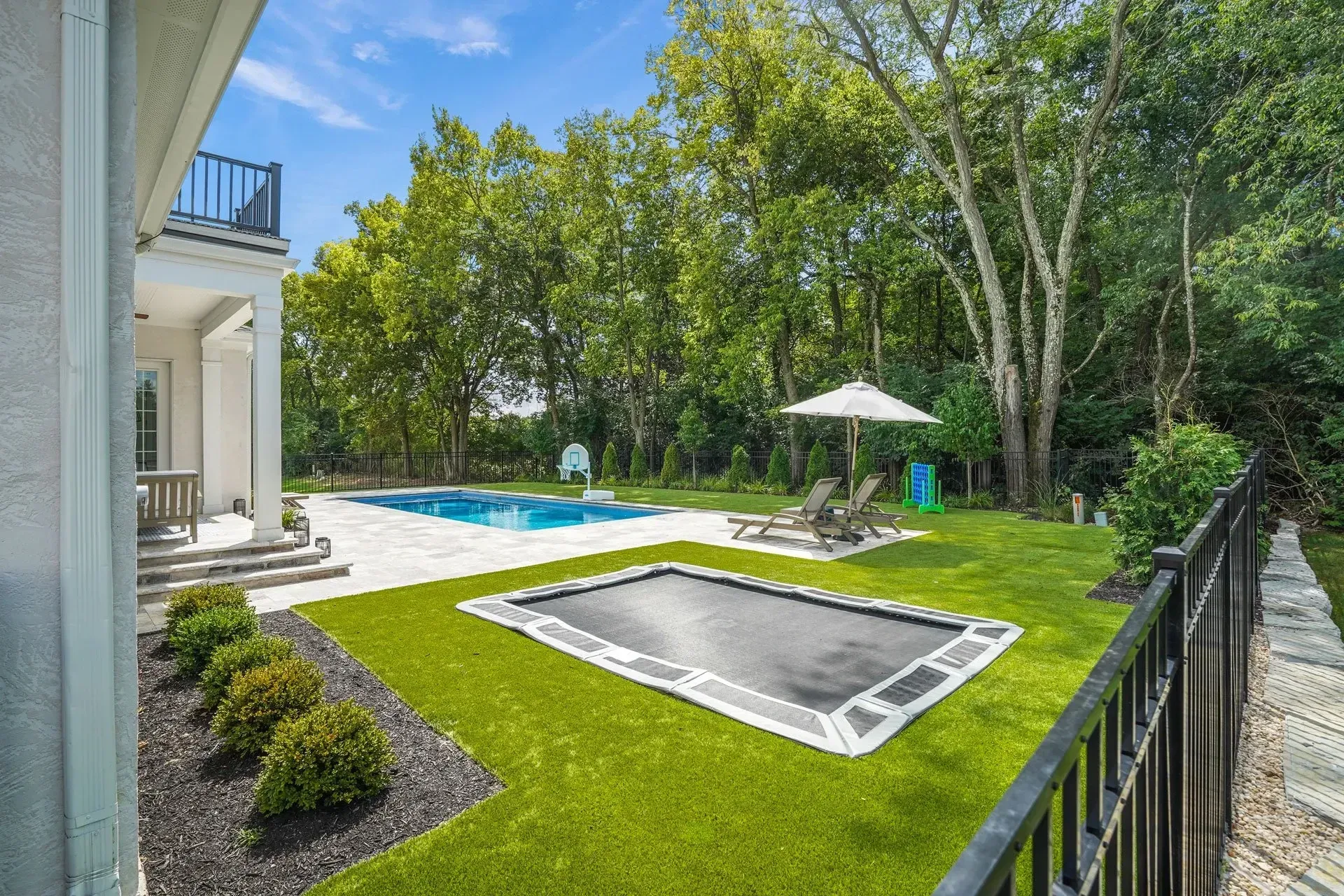 Backyard with a pool, trampoline, and lounge chairs on a sunny day. Green lawn, trees, and a house visible.
