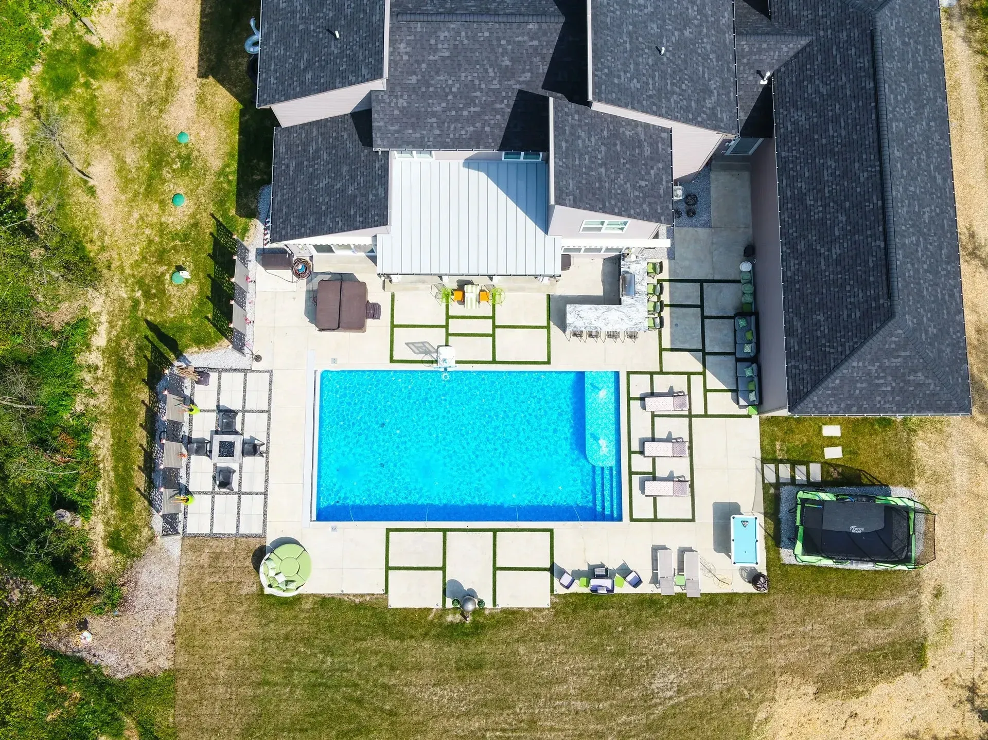 Overhead view of a luxury home with a rectangular pool, patio, and grassy lawn. The roof is dark gray, and the pool water is bright blue.