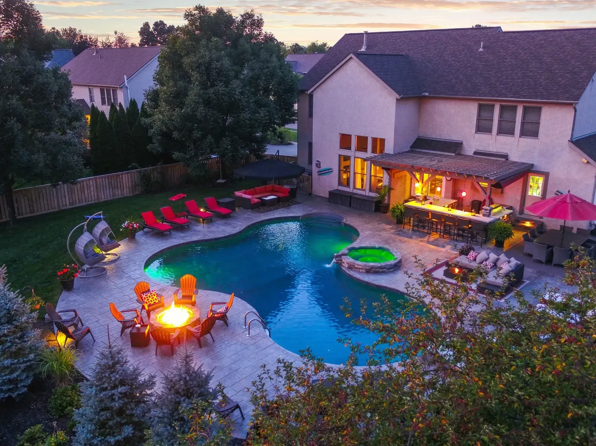 Backyard evening scene with a pool, fire pit, bar, and lounge chairs, lit with ambient lighting.
