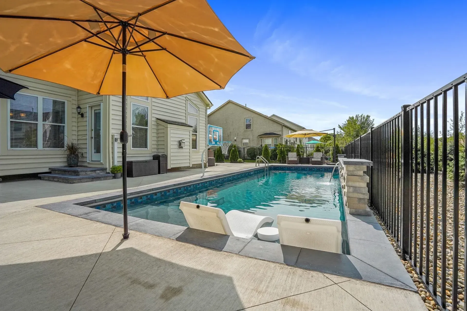 A backyard pool with lounge chairs under an umbrella.  The pool is lined with grey concrete and is surrounded by a black fence and beige house.