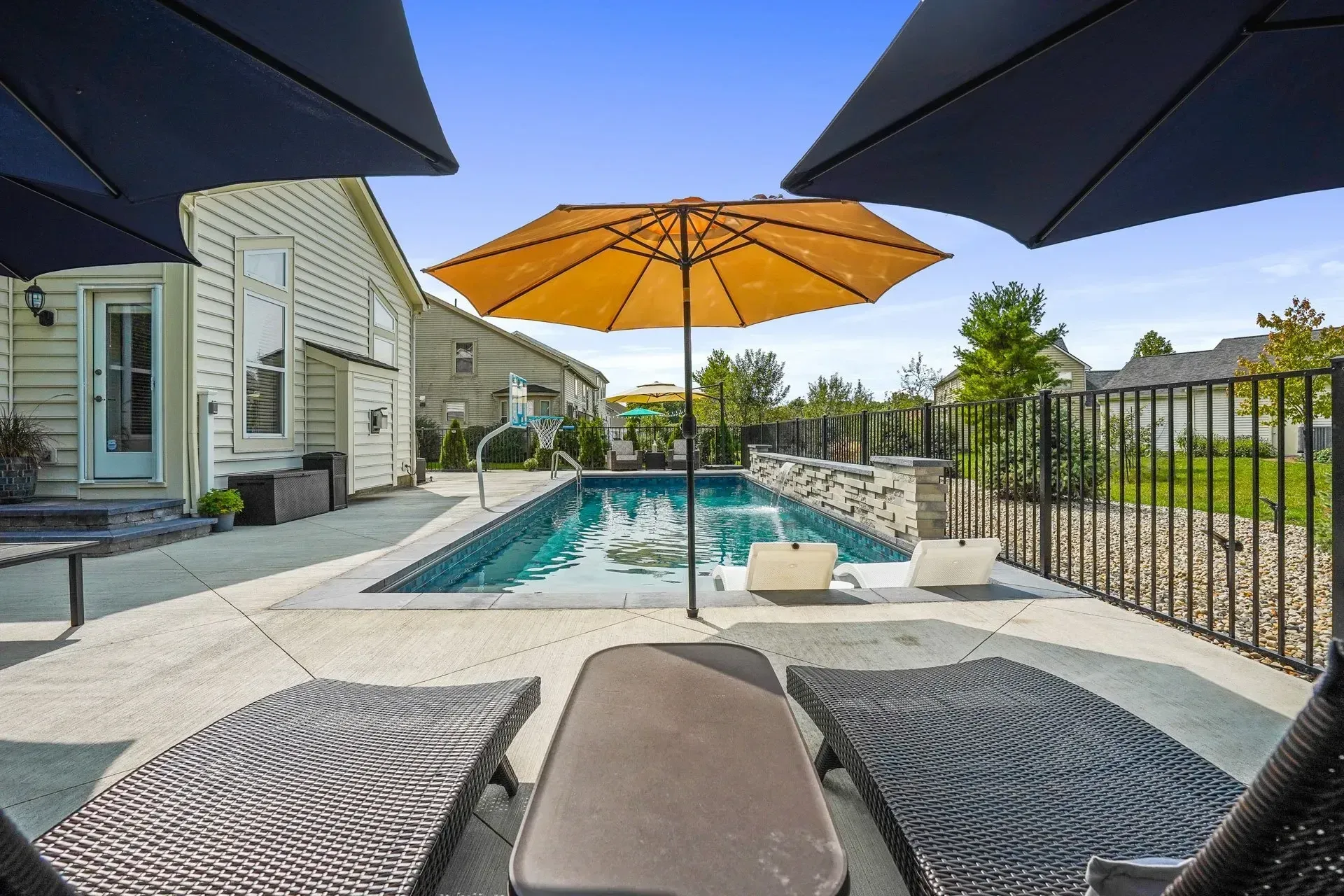 A backyard with a rectangular pool, lounge chairs, and umbrellas on a sunny day. A house is visible in the background.