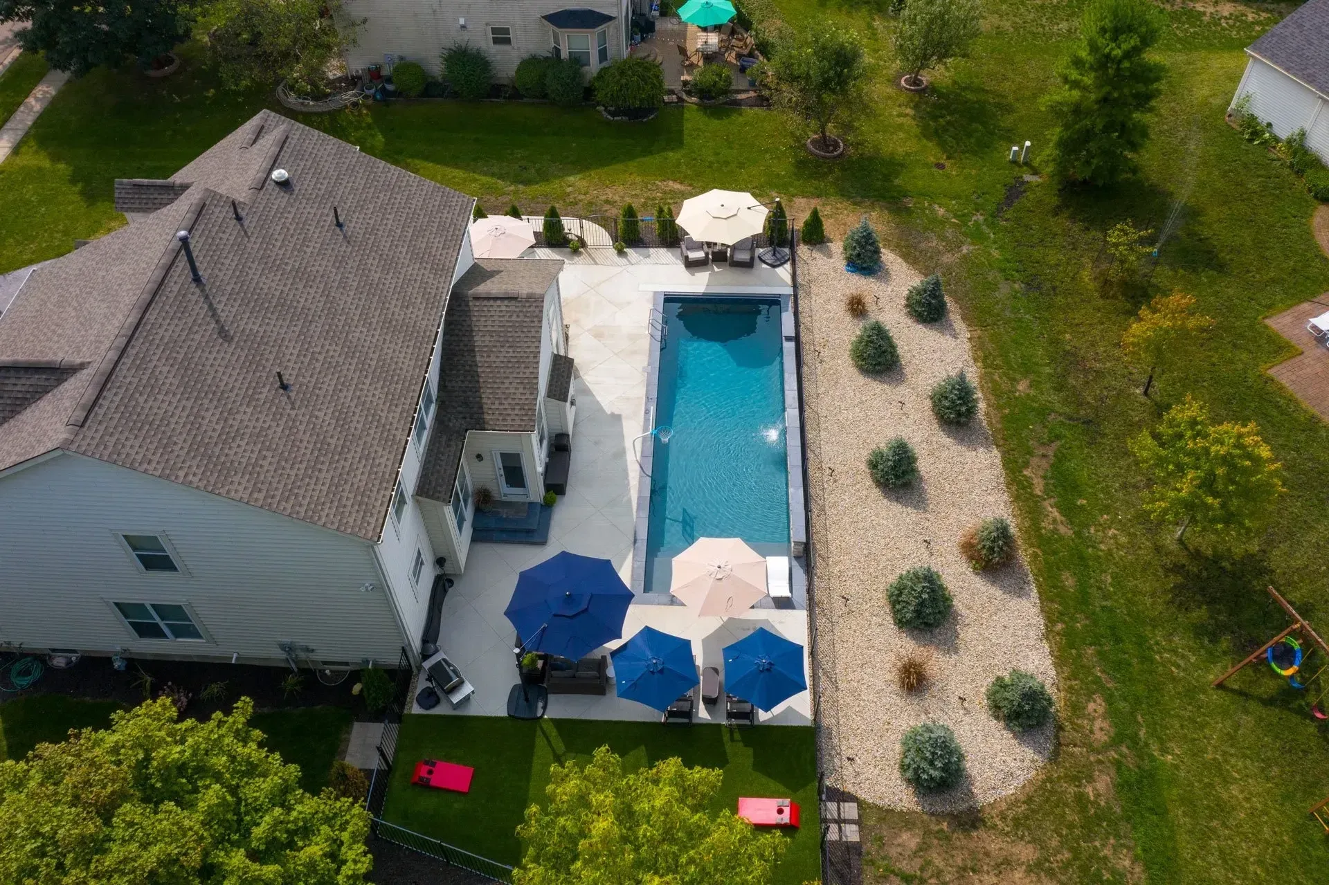 Aerial view of a house with a rectangular swimming pool, patio, and manicured lawn. Umbrellas provide shade near the pool.