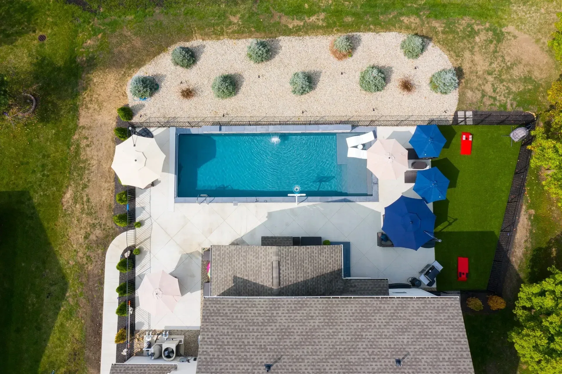 Aerial view of a backyard with a rectangular pool, patio, lawn, and house. Features include umbrellas, lounge chairs, and a decorative rock garden.