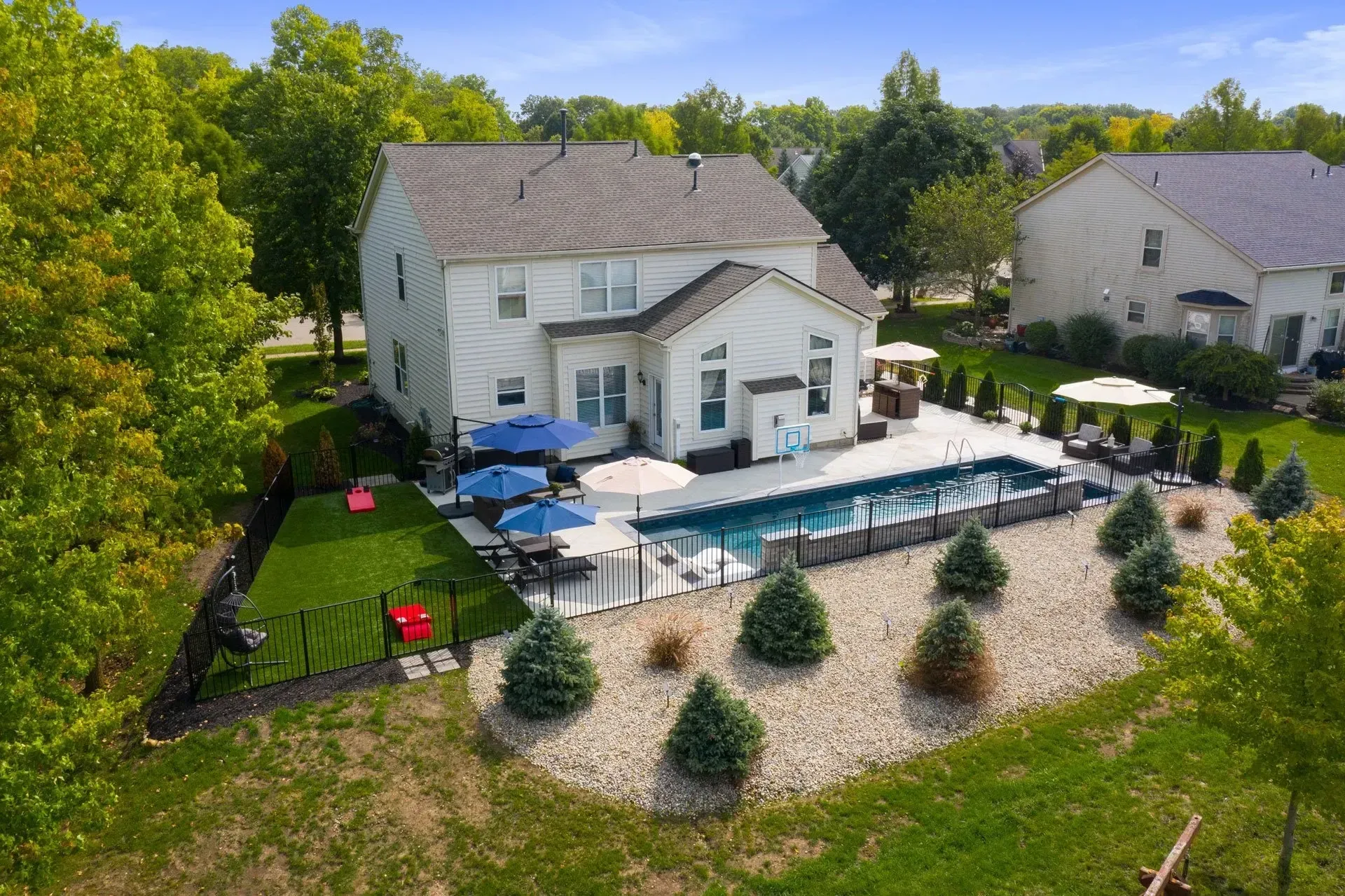 Backyard with a pool, patio, and two-story house. Blue umbrellas shade the patio; evergreen bushes surround the pool.