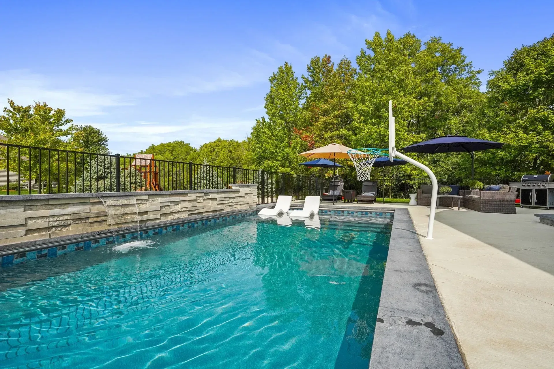 Swimming pool with turquoise water. Features include pool floats, a basketball hoop, and a patio with trees in the background.