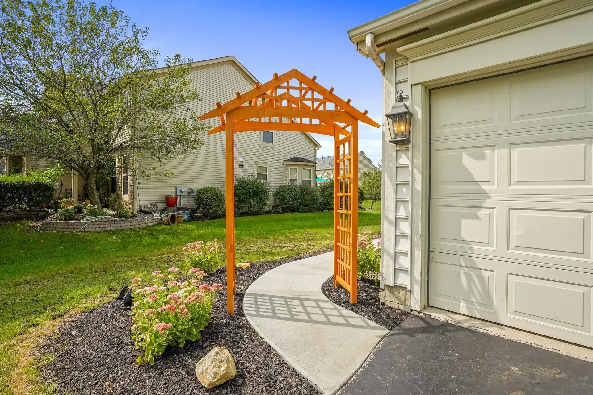 Wooden garden arbor over a curved walkway next to a garage.  Flowers in a mulch bed accent the path.