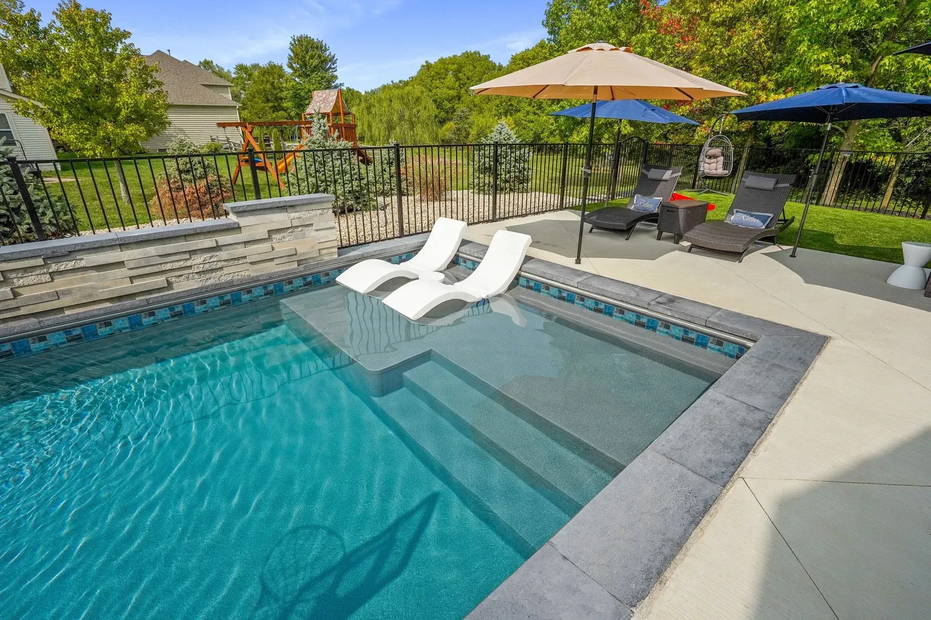 A backyard swimming pool with lounge chairs in the water and on the deck, shaded by umbrellas. Lush green trees surround the pool.
