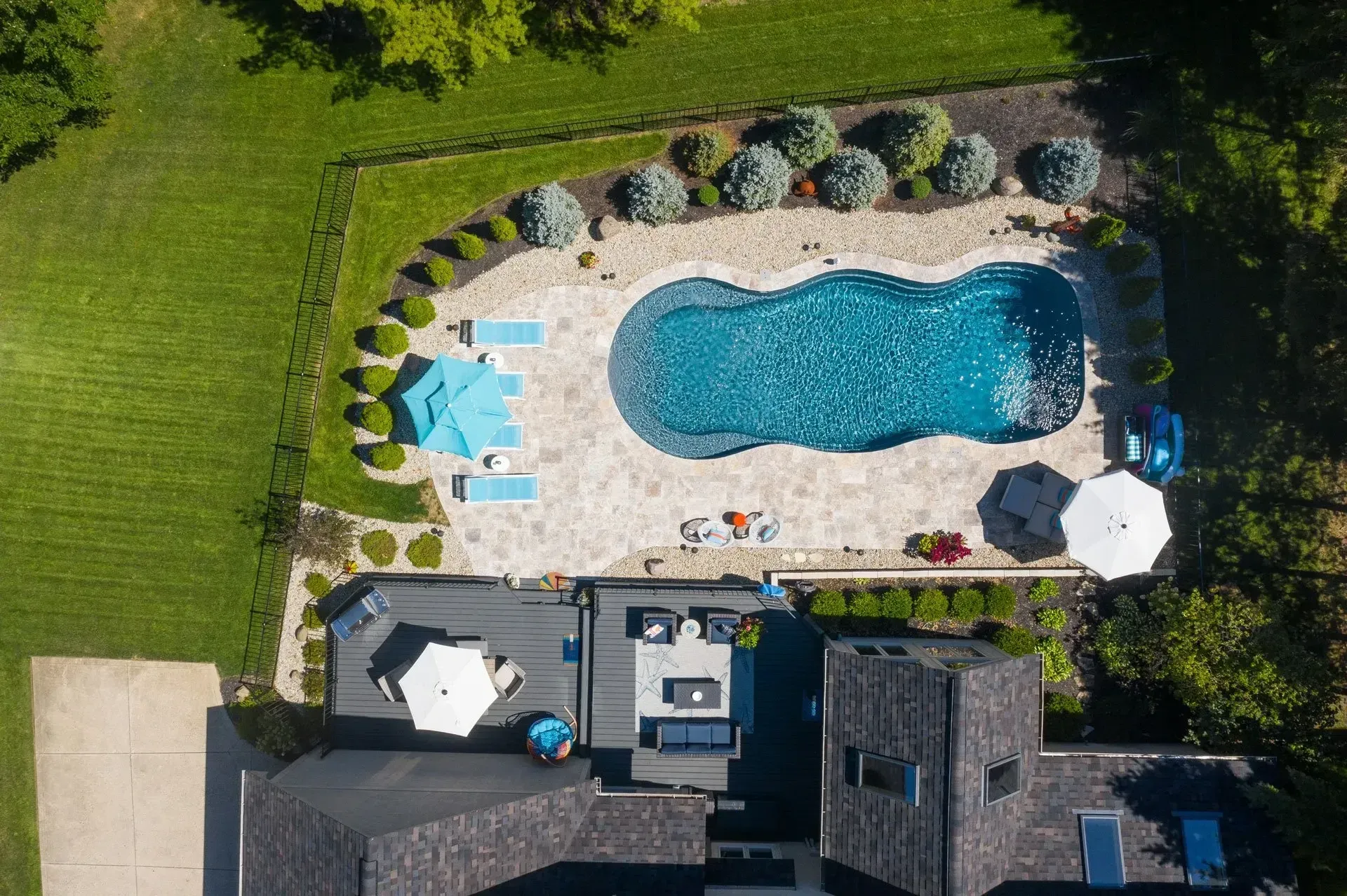 Aerial view of a backyard with a freeform pool, patio, lounge chairs, and landscaping, near a house with a dark roof.