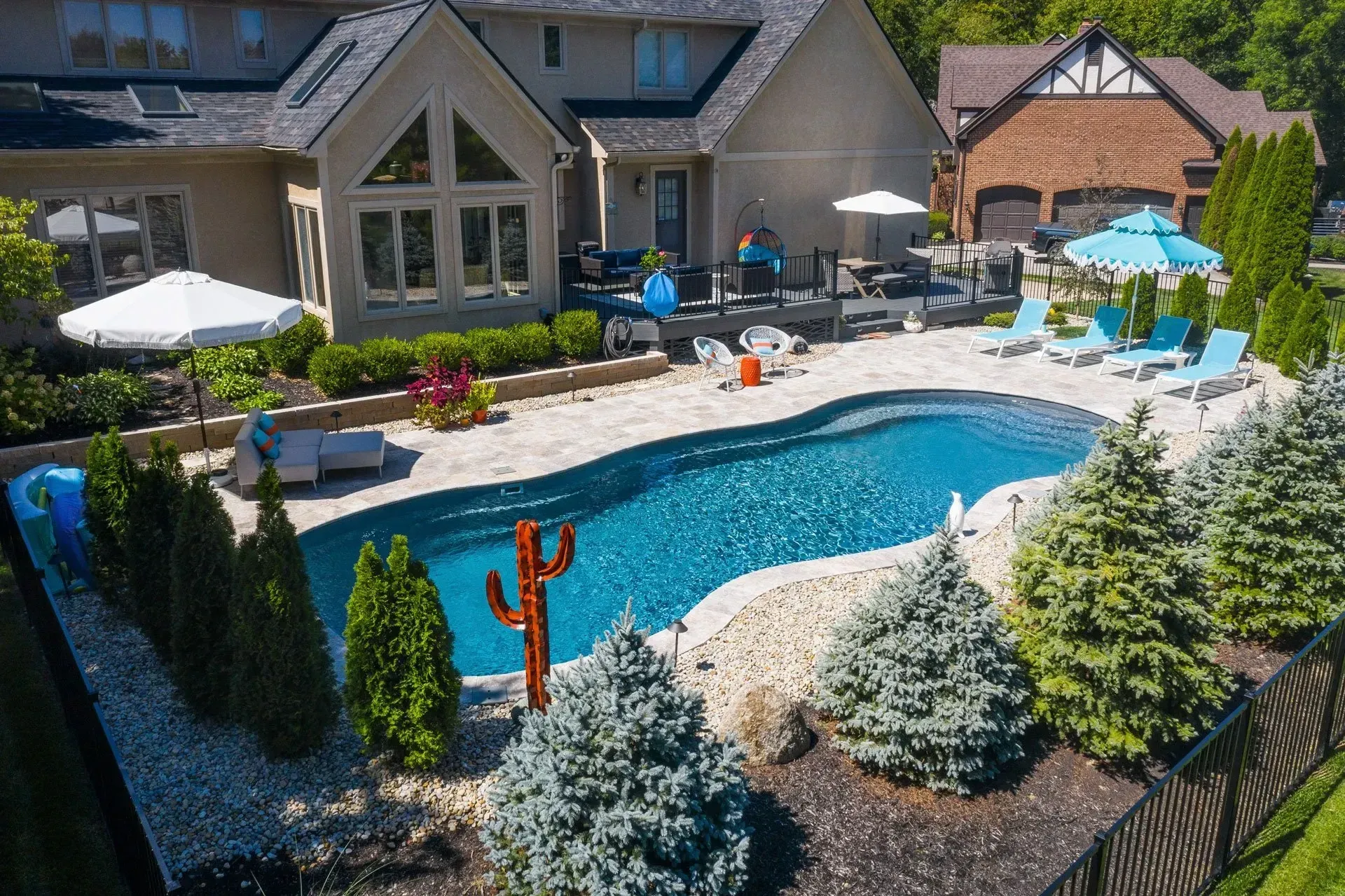 A backyard pool with a house in the background, surrounded by landscaping and patio furniture. Blue water, trees, and a decorative cactus.