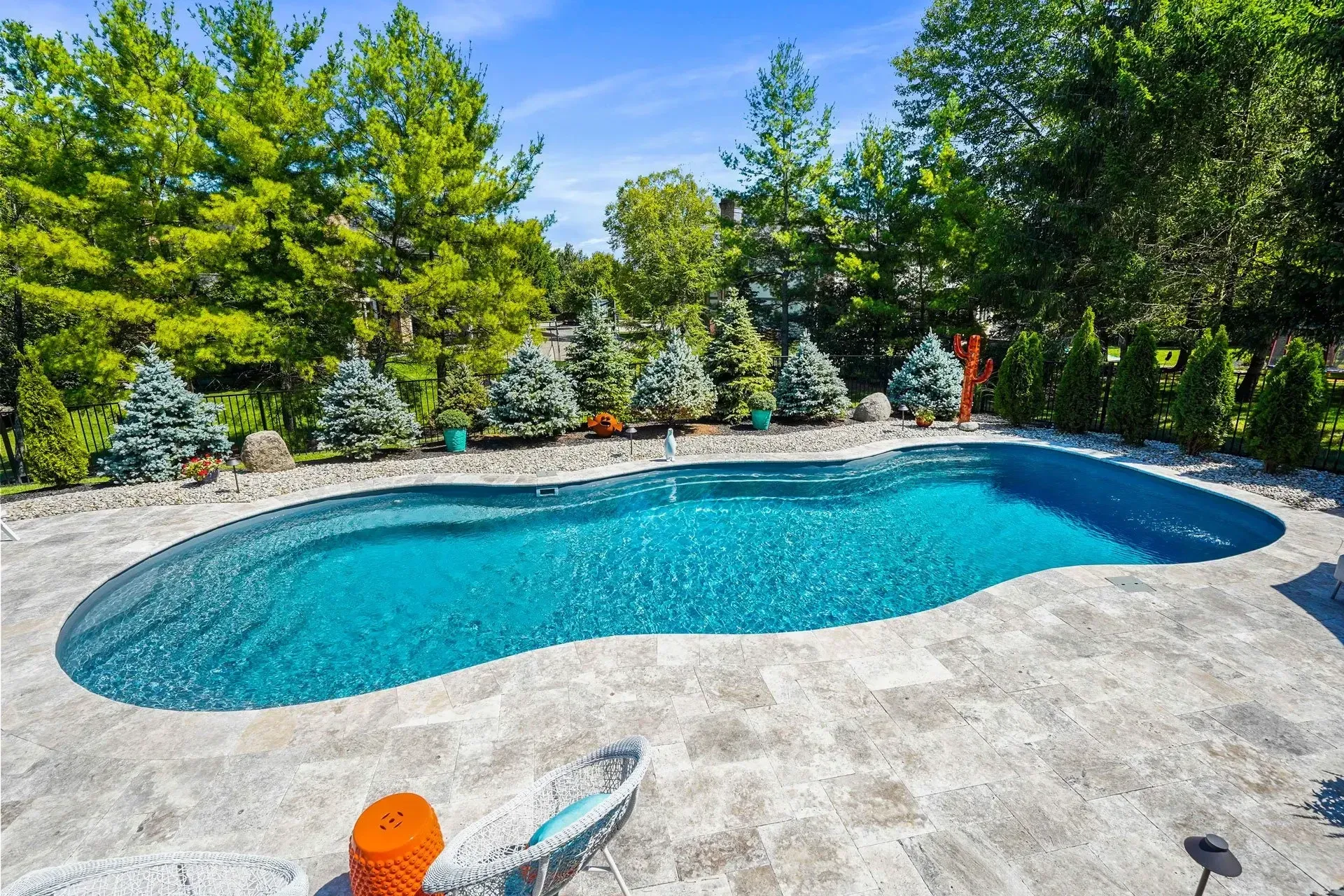 A backyard swimming pool with blue water surrounded by a stone patio and trees on a sunny day.