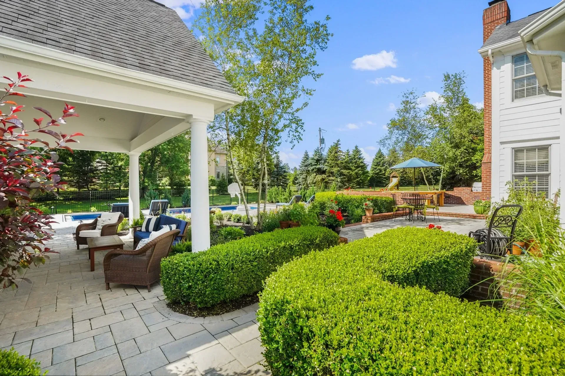 Patio with box hedges, seating area under a white pergola, and a swimming pool. Lush landscaping and a sunny day.