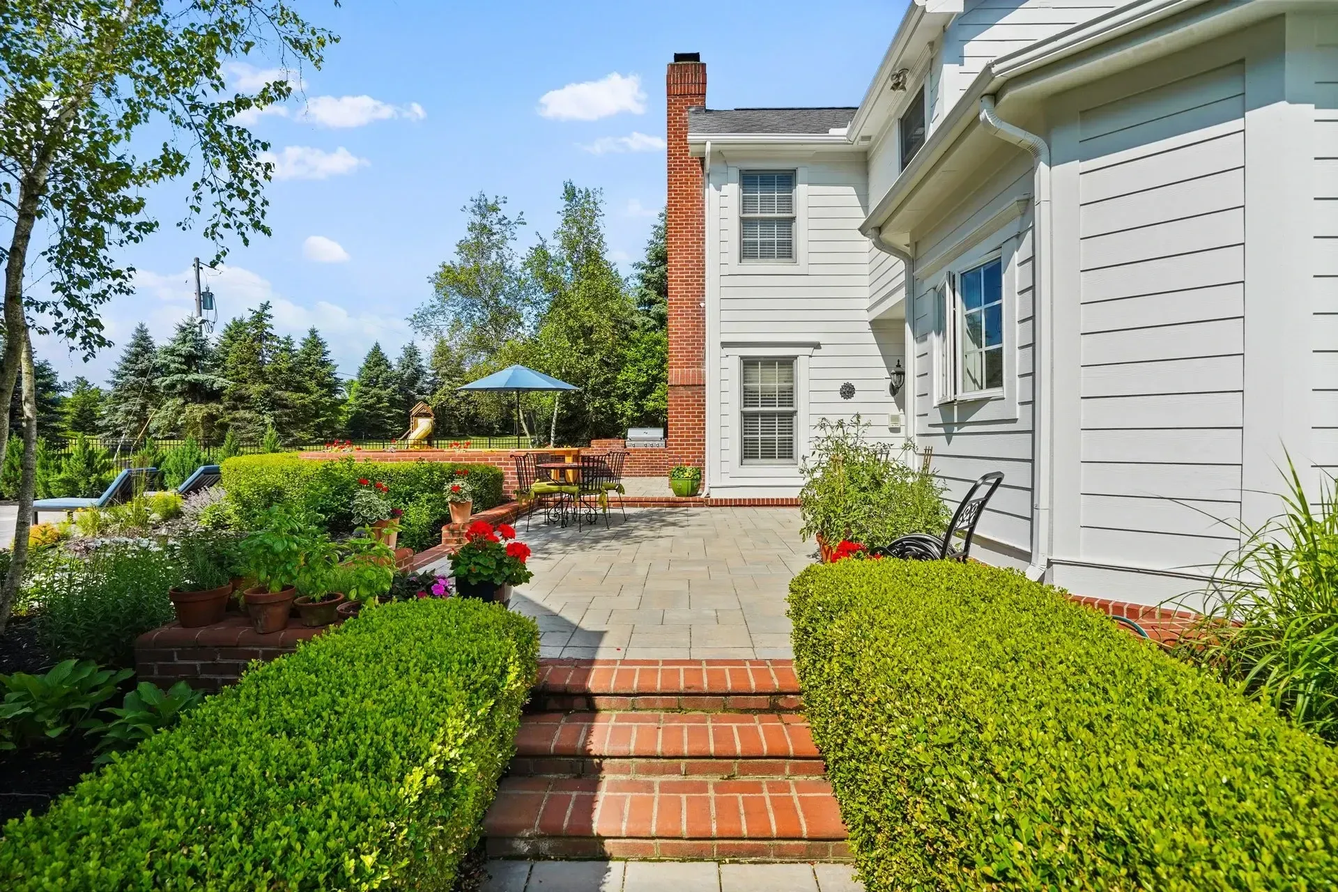 Brick patio and steps lead to a garden with hedges, trees, and a house with a brick chimney.