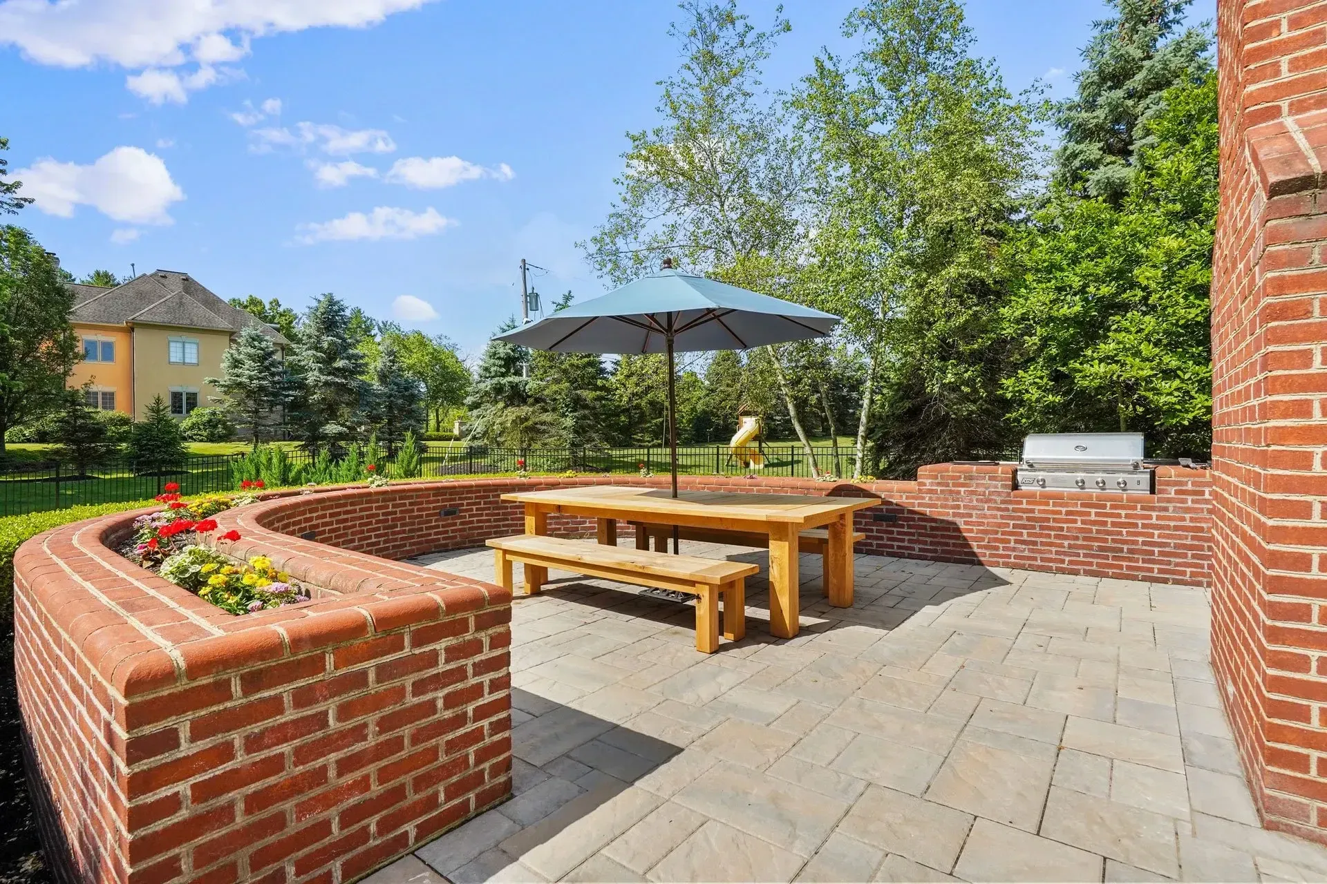 Outdoor patio with brick walls, wooden picnic table, umbrella, and a built-in grill, surrounded by green trees and lawn, sunny day.