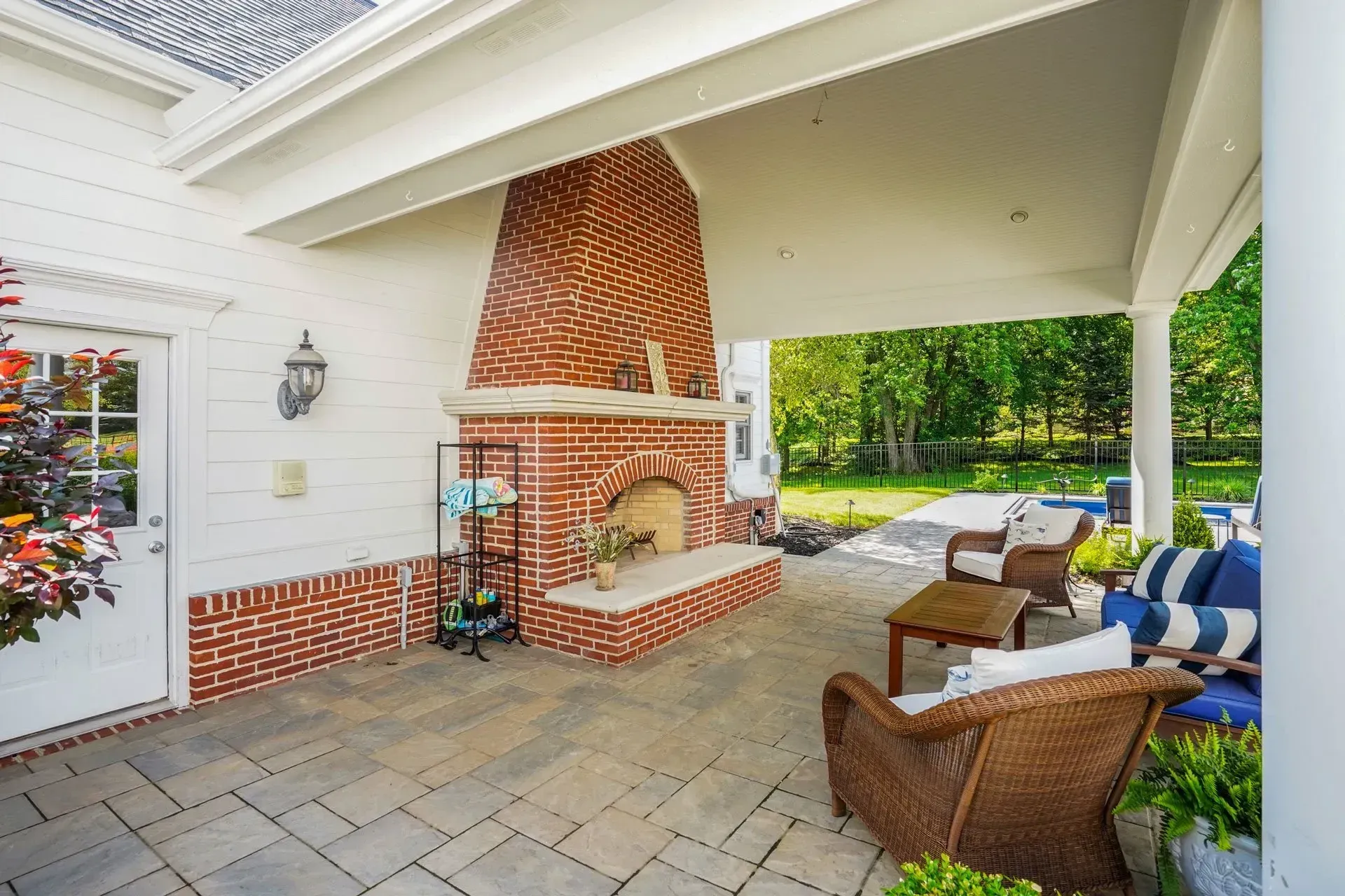 Covered patio with brick fireplace, seating, and view of a green yard. White columns and trim.