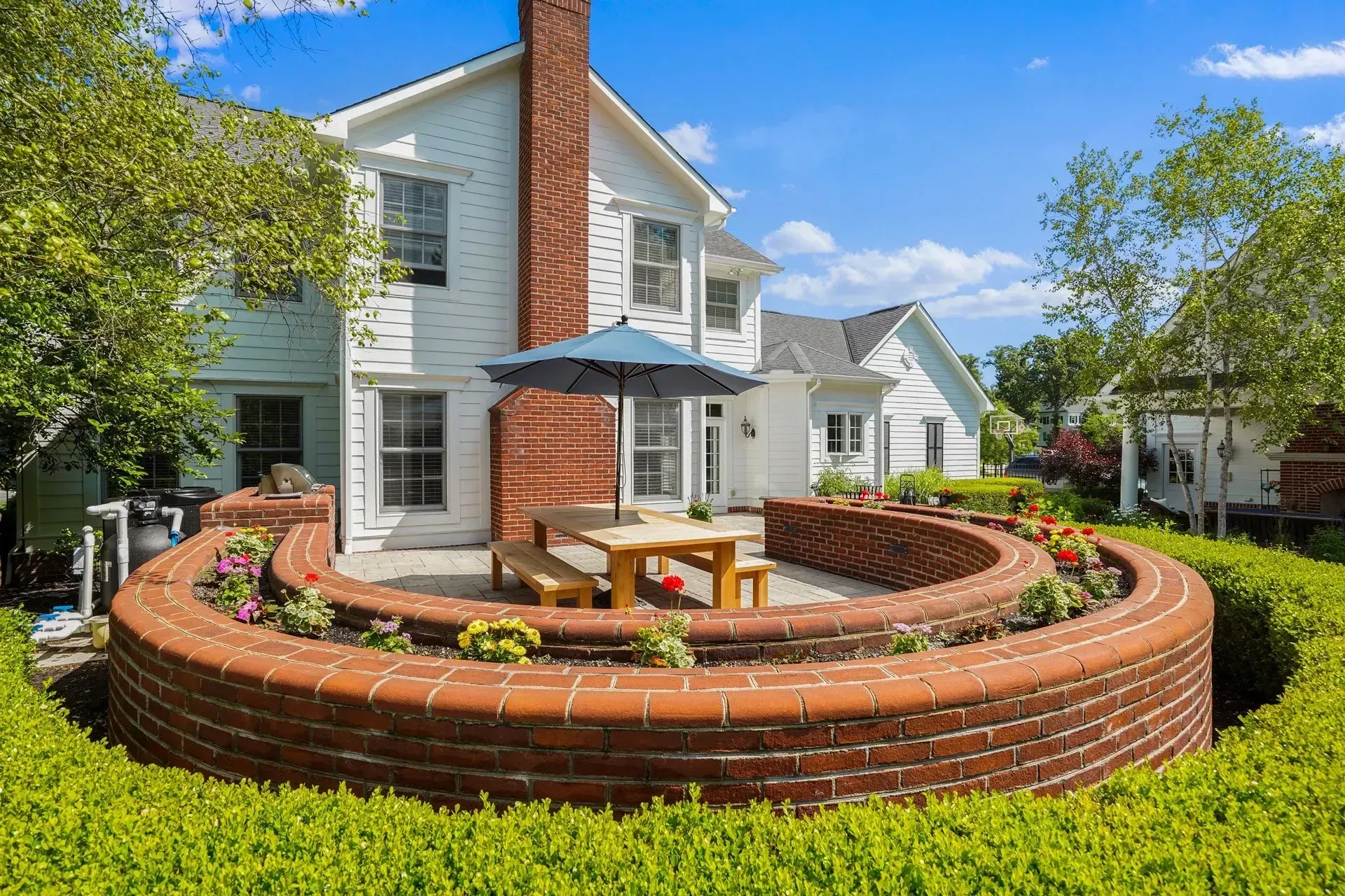 White house with a brick-walled patio, outdoor table, and floral gardens, surrounded by neatly trimmed hedges.