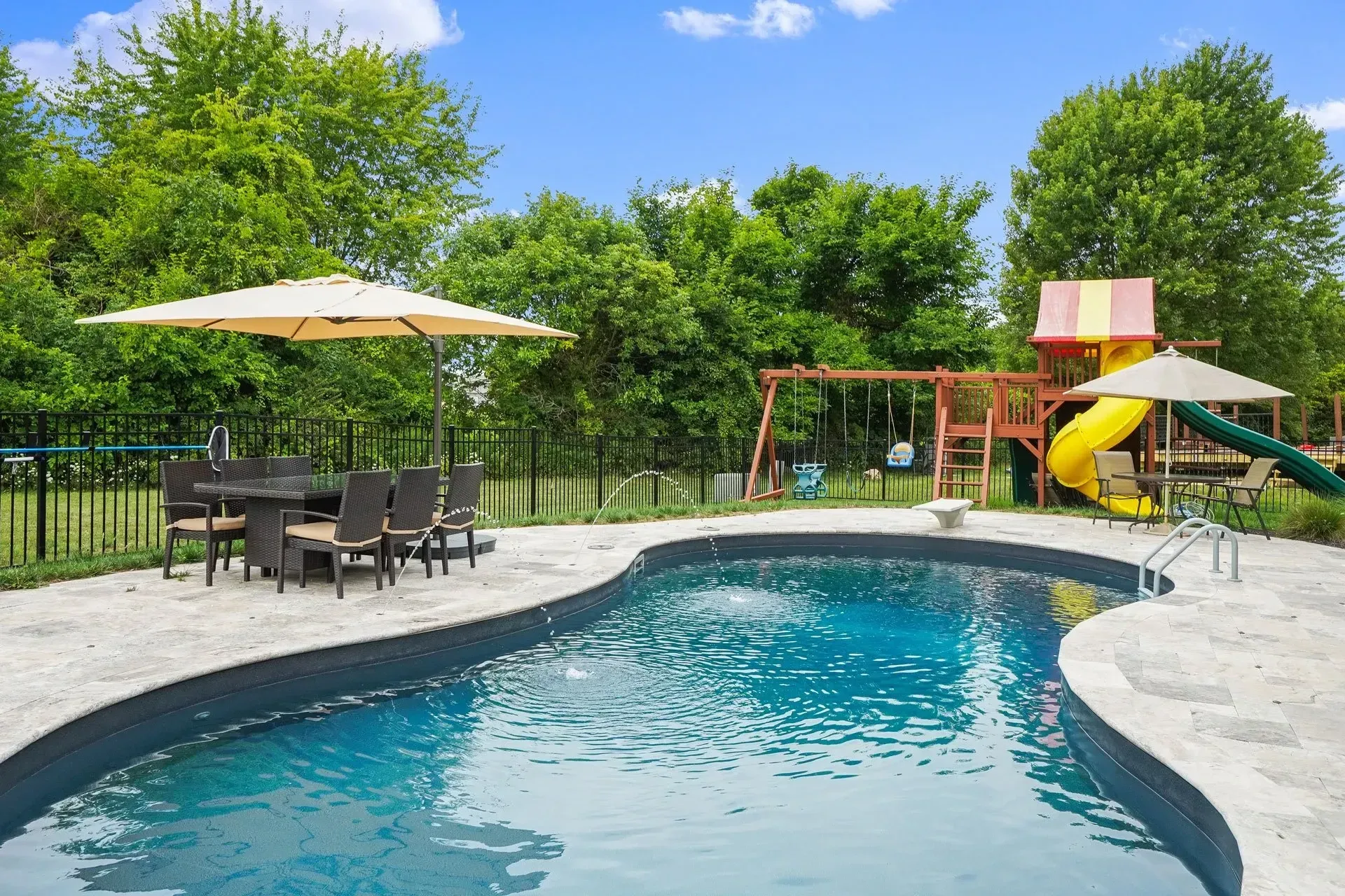 A backyard pool with a dining area, a playground, and a clear blue sky. The pool is surrounded by light-colored concrete.