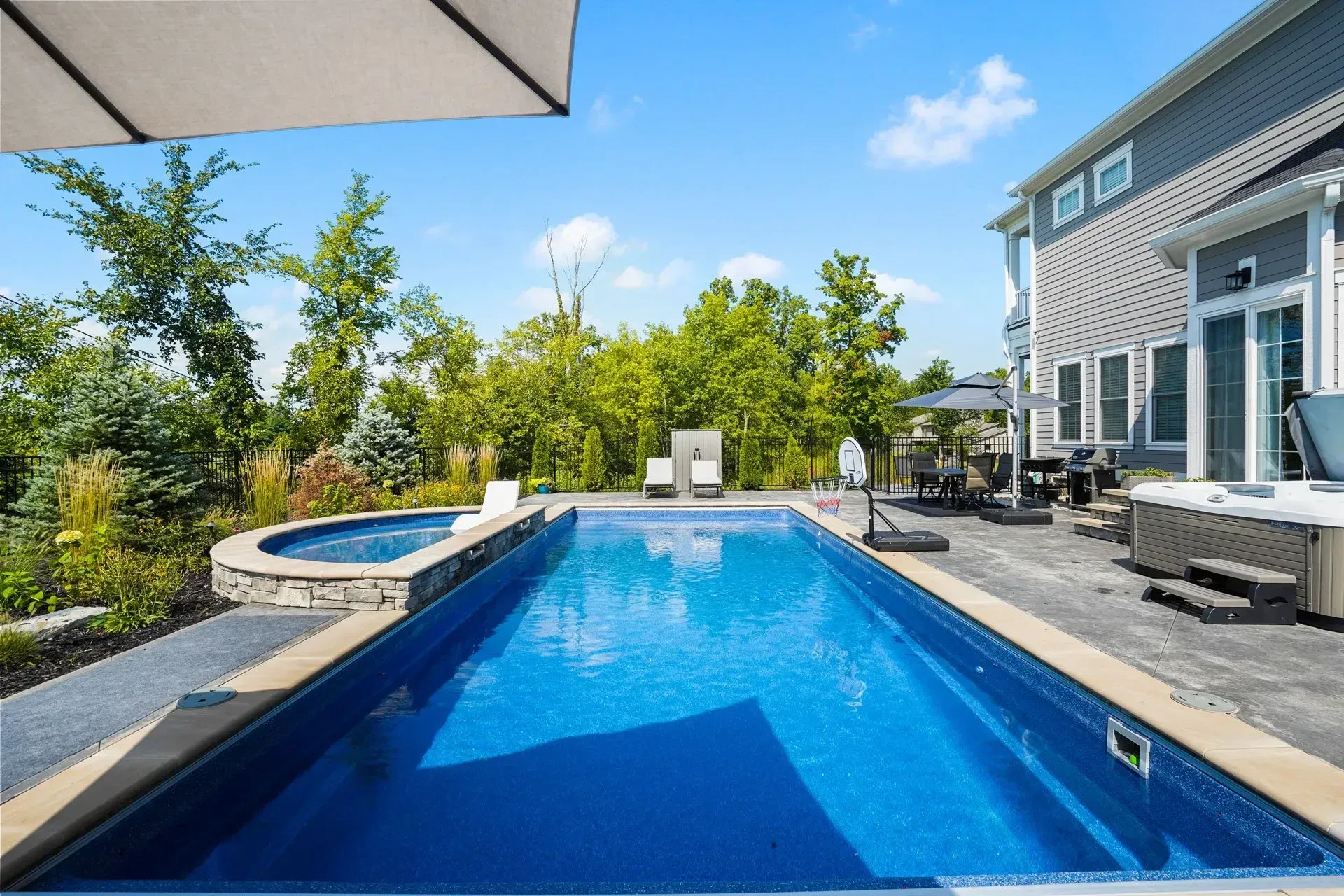 Backyard pool with a spa, surrounded by a patio and a house. Blue water reflects the sunny sky.