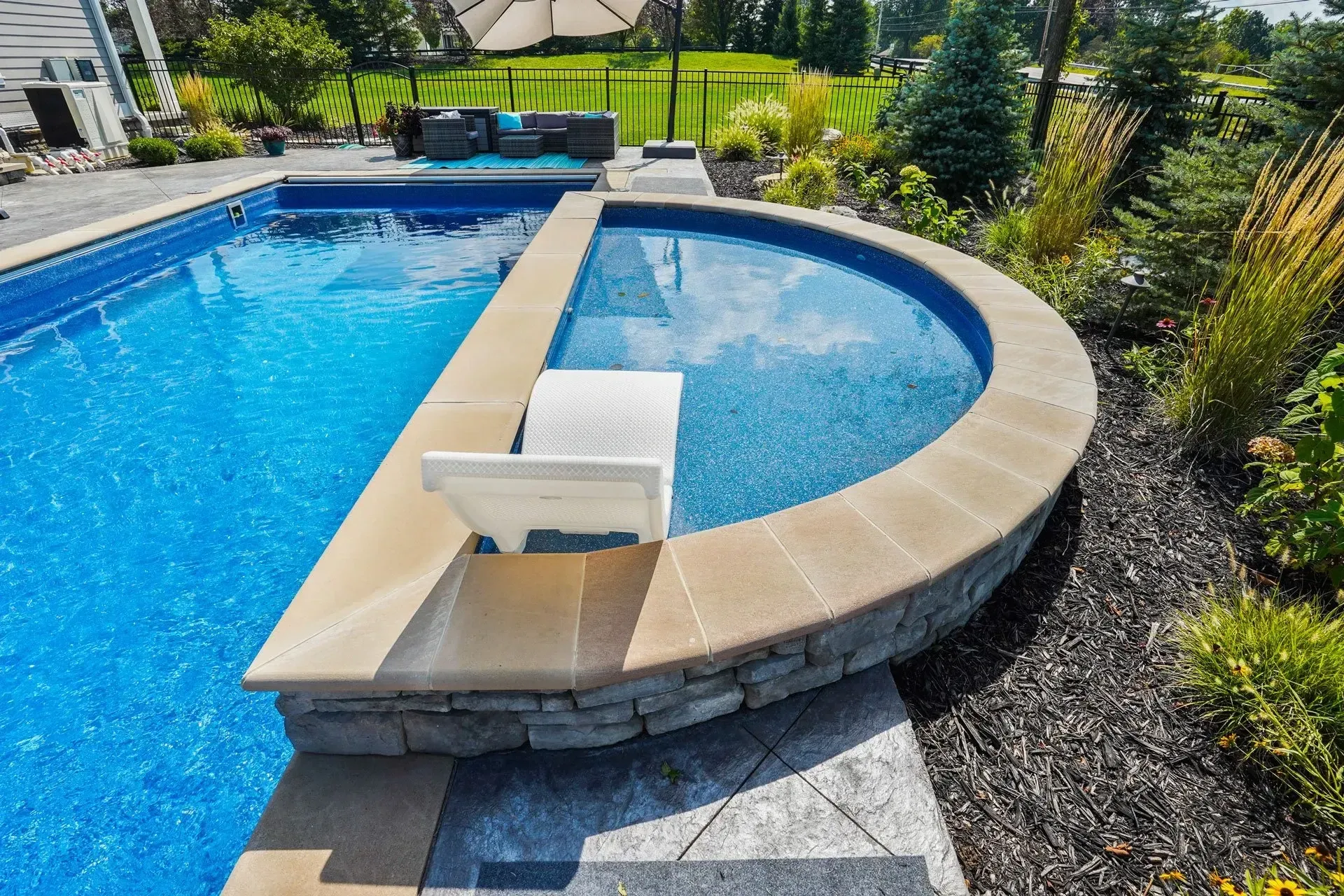 A backyard pool with a built-in spa. Blue water, stone surround, and landscaping with a sunny background.