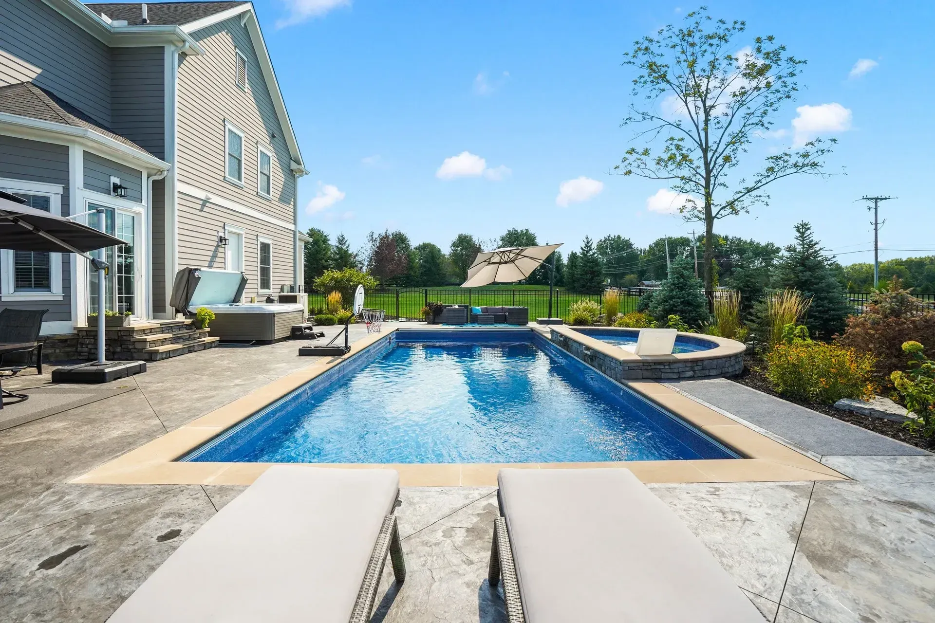 Backyard with a rectangular swimming pool, hot tub, and lounging chairs in front of a gray house under a bright blue sky.