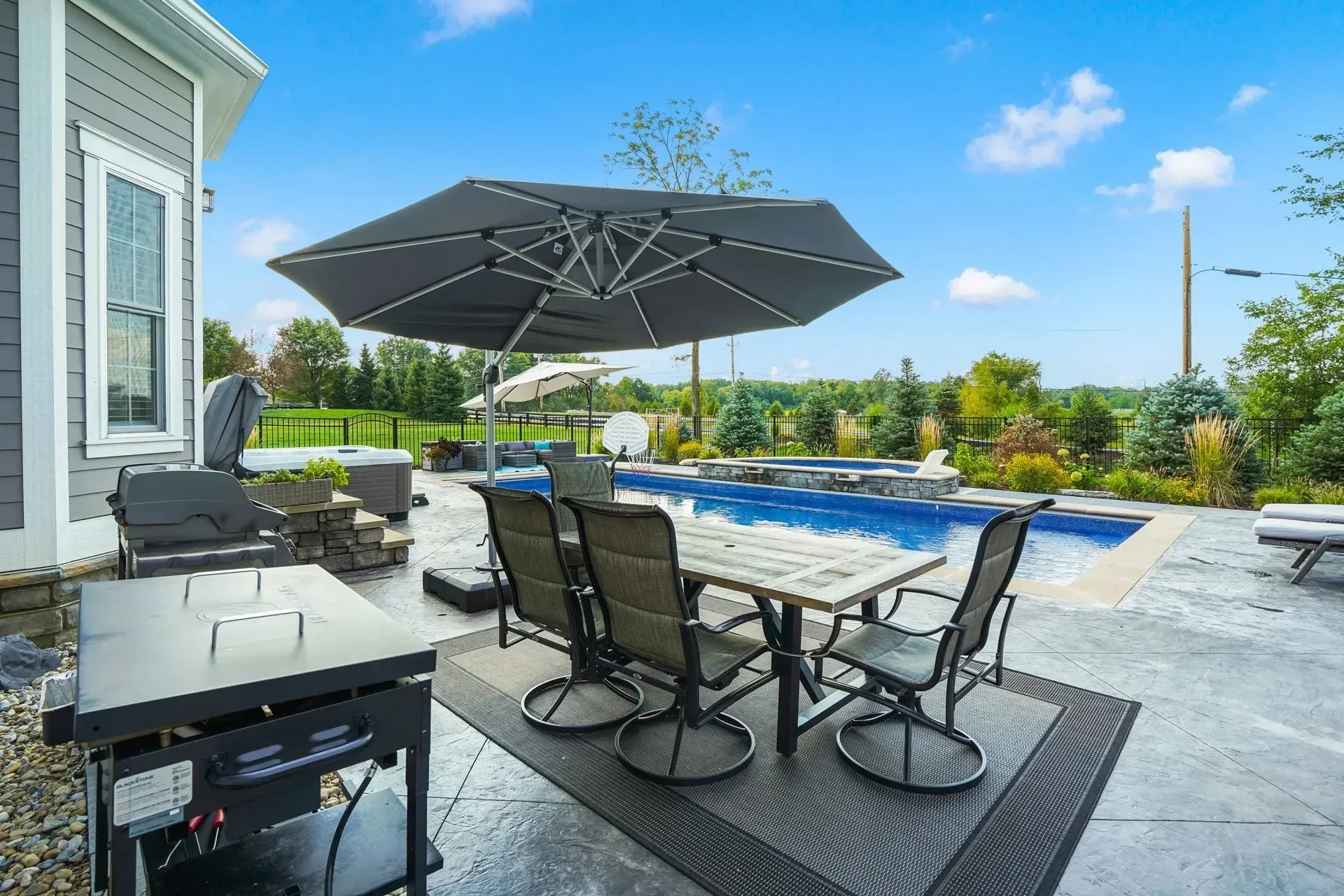 Backyard patio with a pool, grill, dining table under an umbrella, and a clear blue sky.