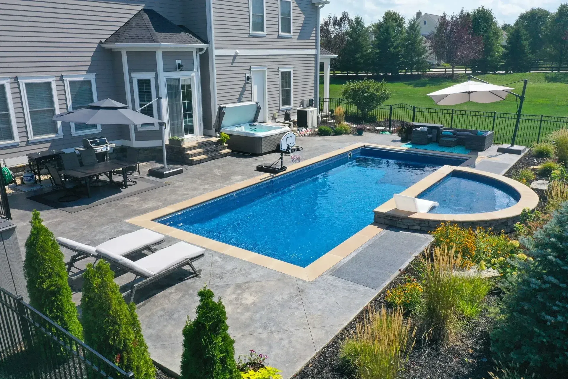 Backyard with a rectangular pool, hot tub, and patio furniture. House in the background with a lawn and trees.