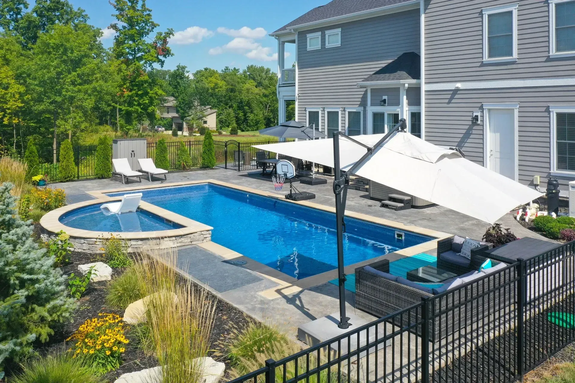 Backyard pool with blue water, next to a house. Features include a spa, lounge chairs, umbrella, and black fence.