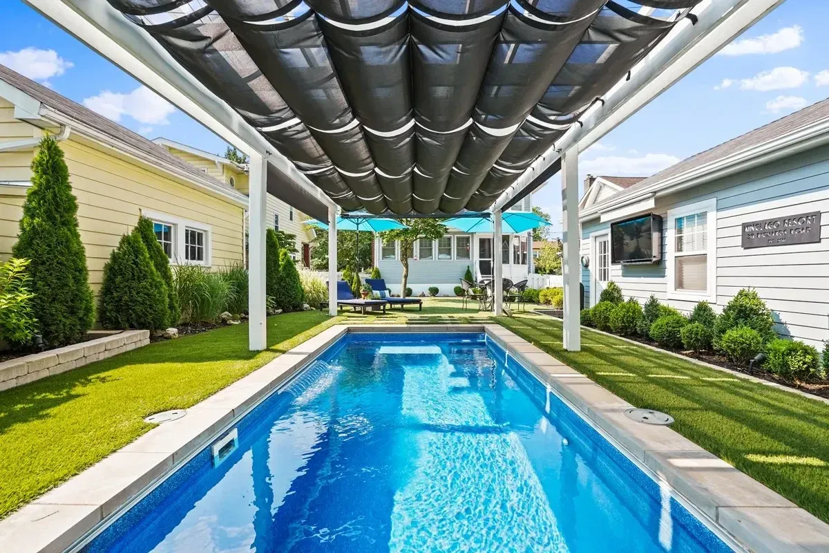 Long, narrow outdoor pool with black retractable shade, set between two homes with a grassy lawn.