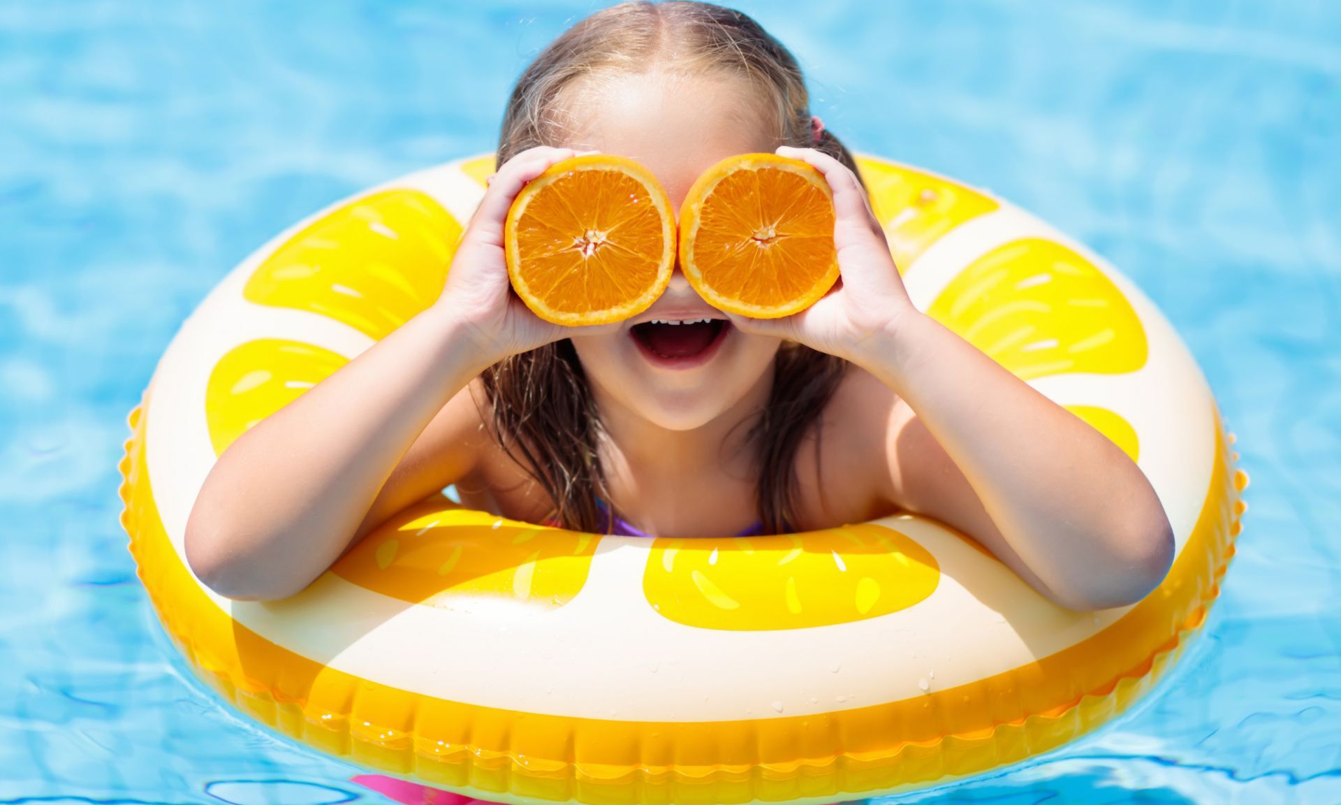 A young girl in a pool, holding an orange slice, while floating on a kid pool floater.