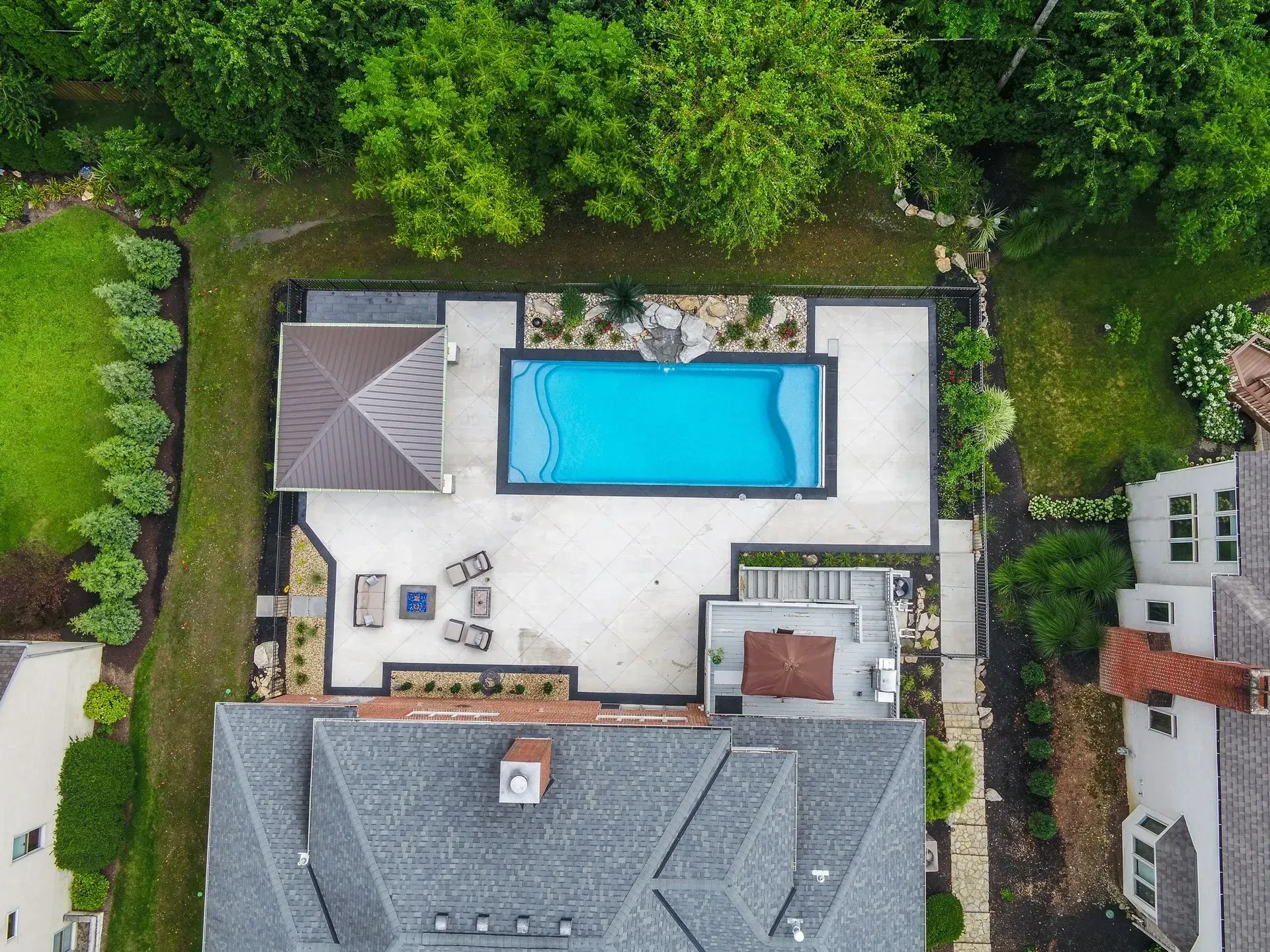 Aerial view of a backyard with a rectangular pool, patio furniture, and a gazebo. Surrounding the pool are stone walkways and lush landscaping.