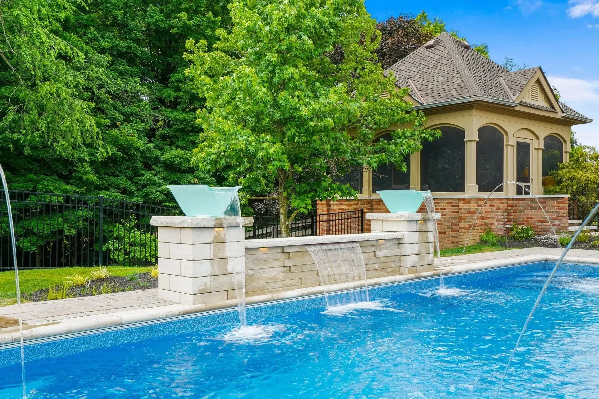 Pool with fountains, a stone wall, and a gazebo in a lush, green setting under a blue sky.