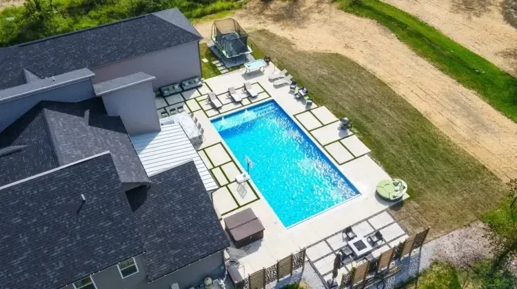 Aerial view of a rectangular swimming pool with a hot tub and lounge chairs in a backyard setting next to a house.
