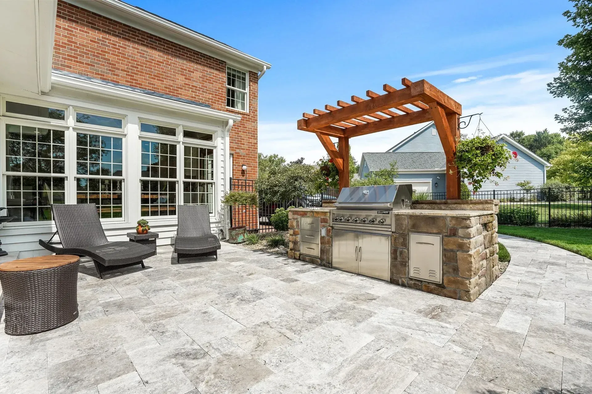 Outdoor patio with built-in grill under a wooden pergola next to a brick house.  Lounge chairs sit near the window.