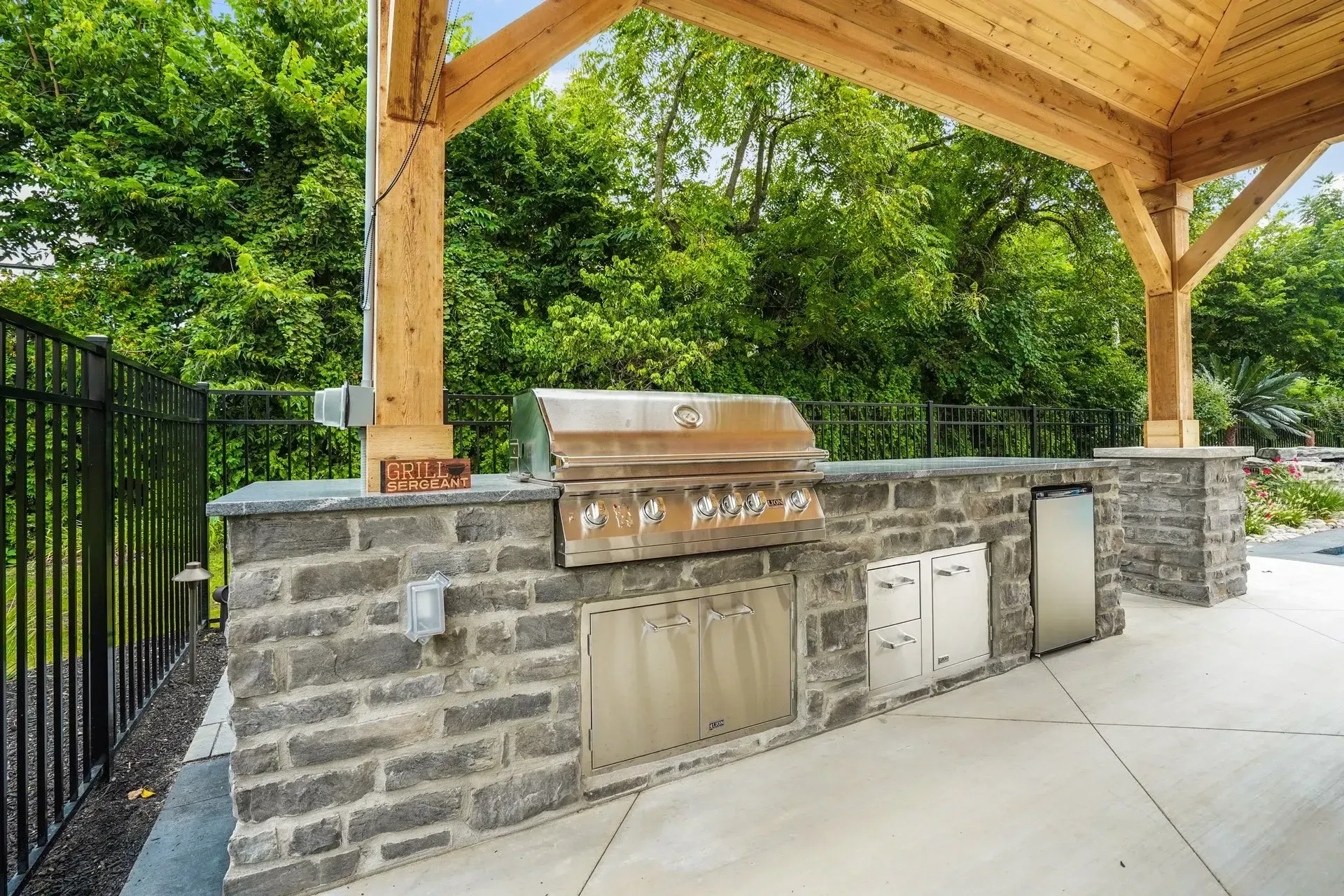 Outdoor kitchen with a grill, storage, and a refrigerator, built into a stone structure under a wooden pergola.