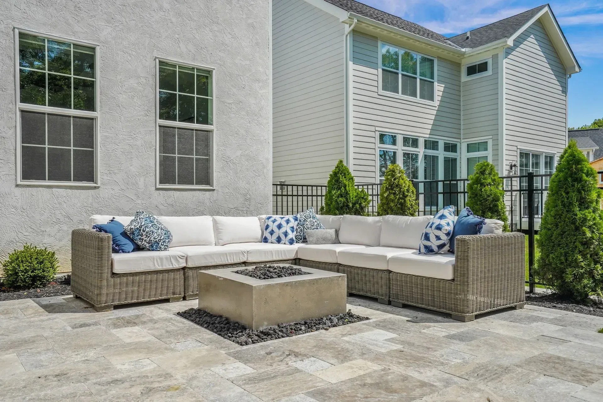 Outdoor patio with sectional seating around a concrete fire pit. Gray stucco building and greenery in the background.