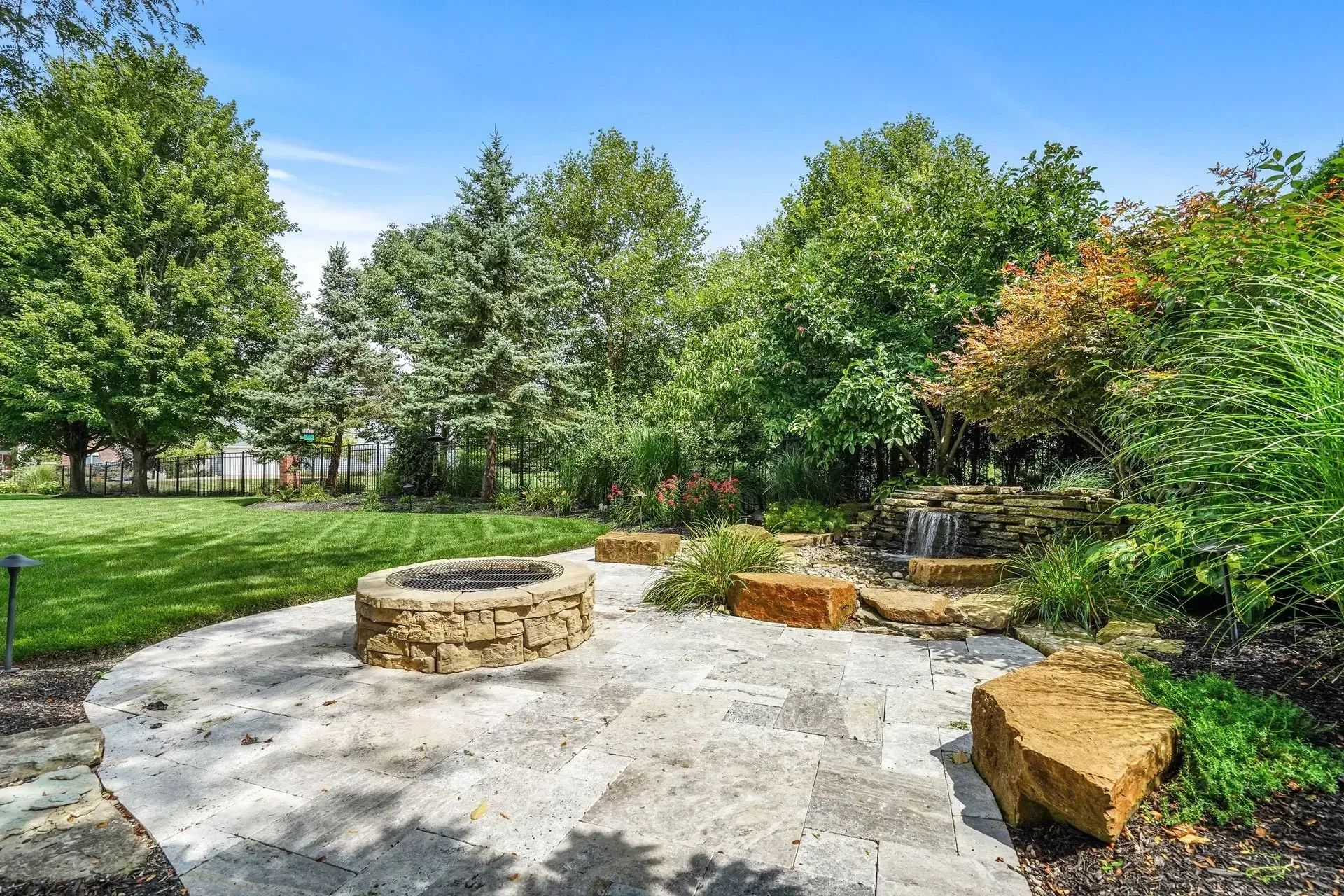 Stone patio with fire pit and small waterfall surrounded by trees and greenery.