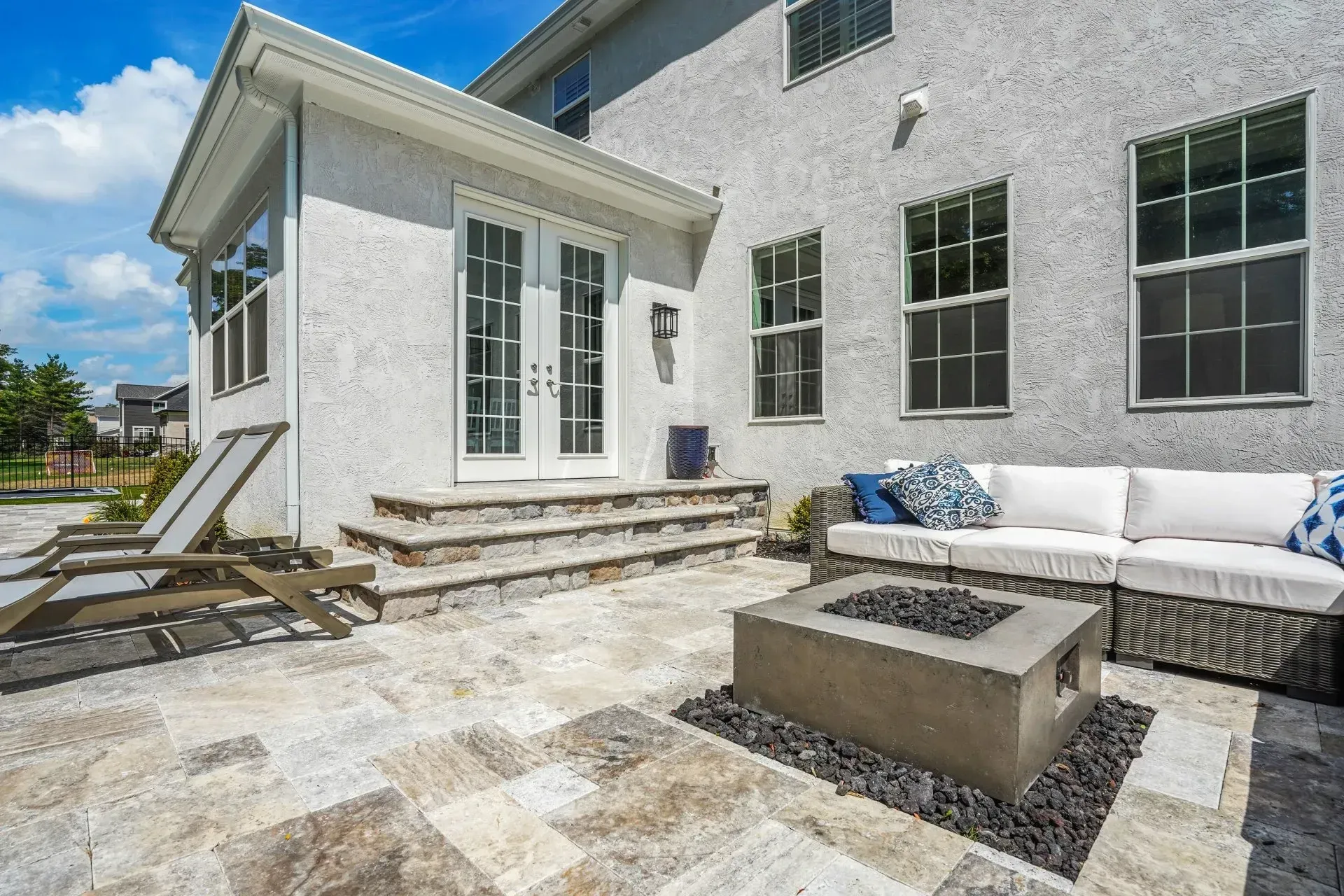 Backyard patio with a grey concrete fire pit, lounge seating, and a stucco house with double doors.