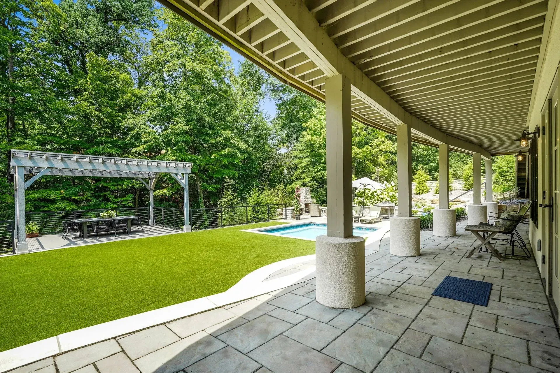 A patio with stone flooring overlooks a green lawn, pool, and pergola.  Green trees create a backdrop under a blue sky.