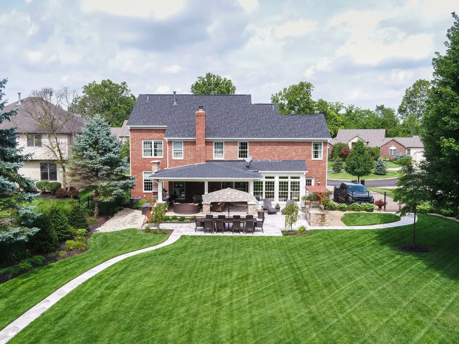 Backyard view of a large brick house with a patio, manicured lawn, and surrounding trees under a cloudy sky.