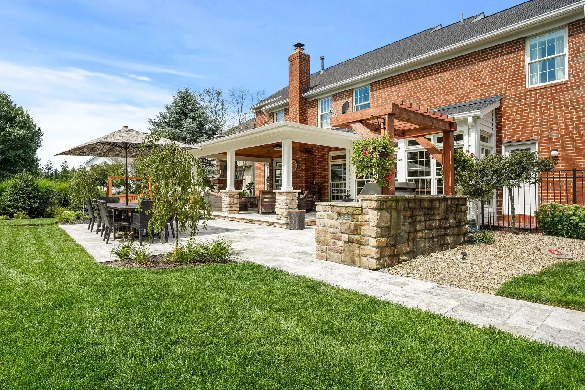A backyard patio with a brick house, stone patio, and green lawn on a sunny day. It features a pergola, dining table, and built-in stone structure.