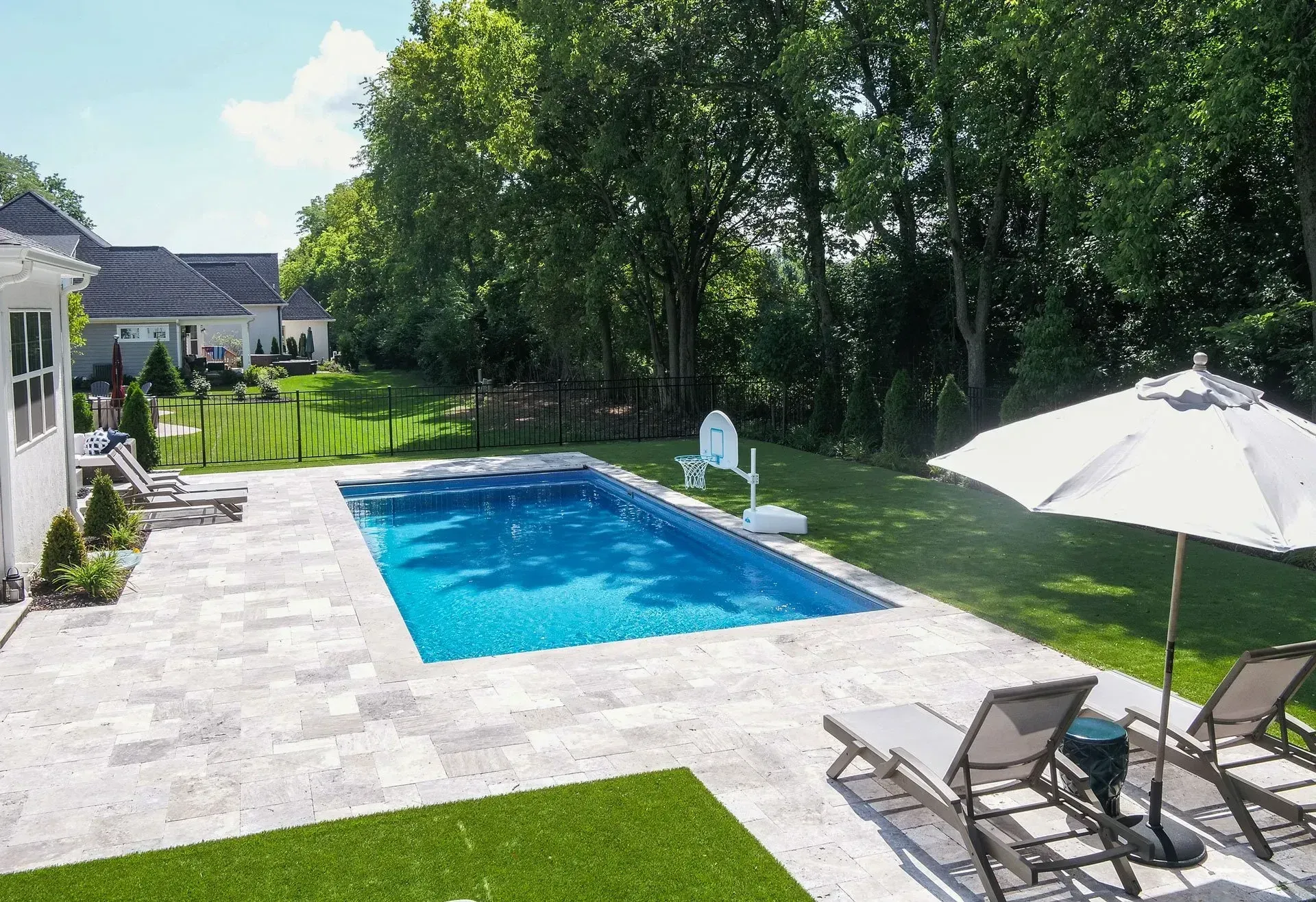 Backyard with rectangular pool, lounge chairs, and umbrella on a stone patio. Green lawn surrounds the pool with trees in the background.
