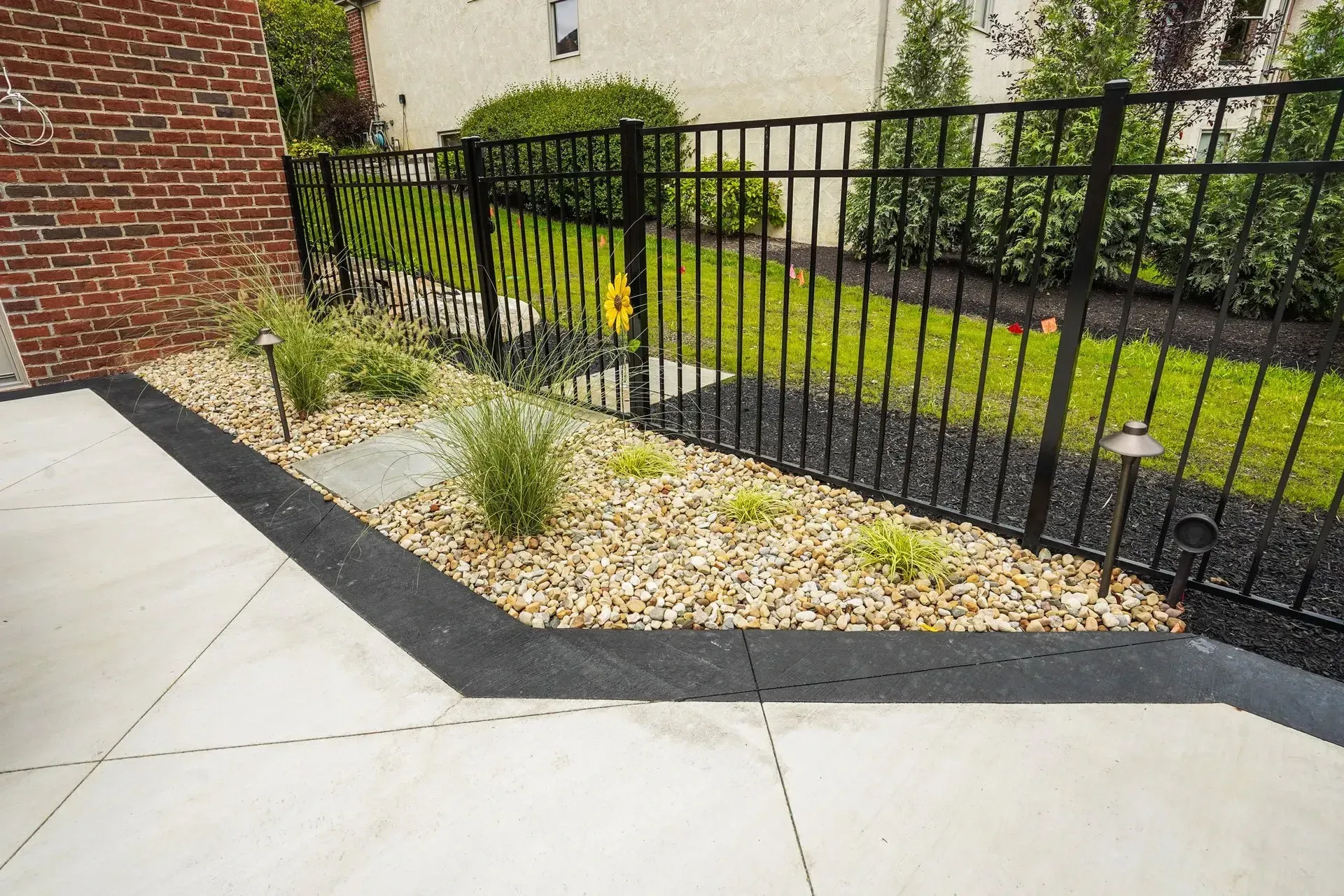 Concrete patio with black trim and a gravel bed next to a black fence. Small plants and lights are in the gravel.