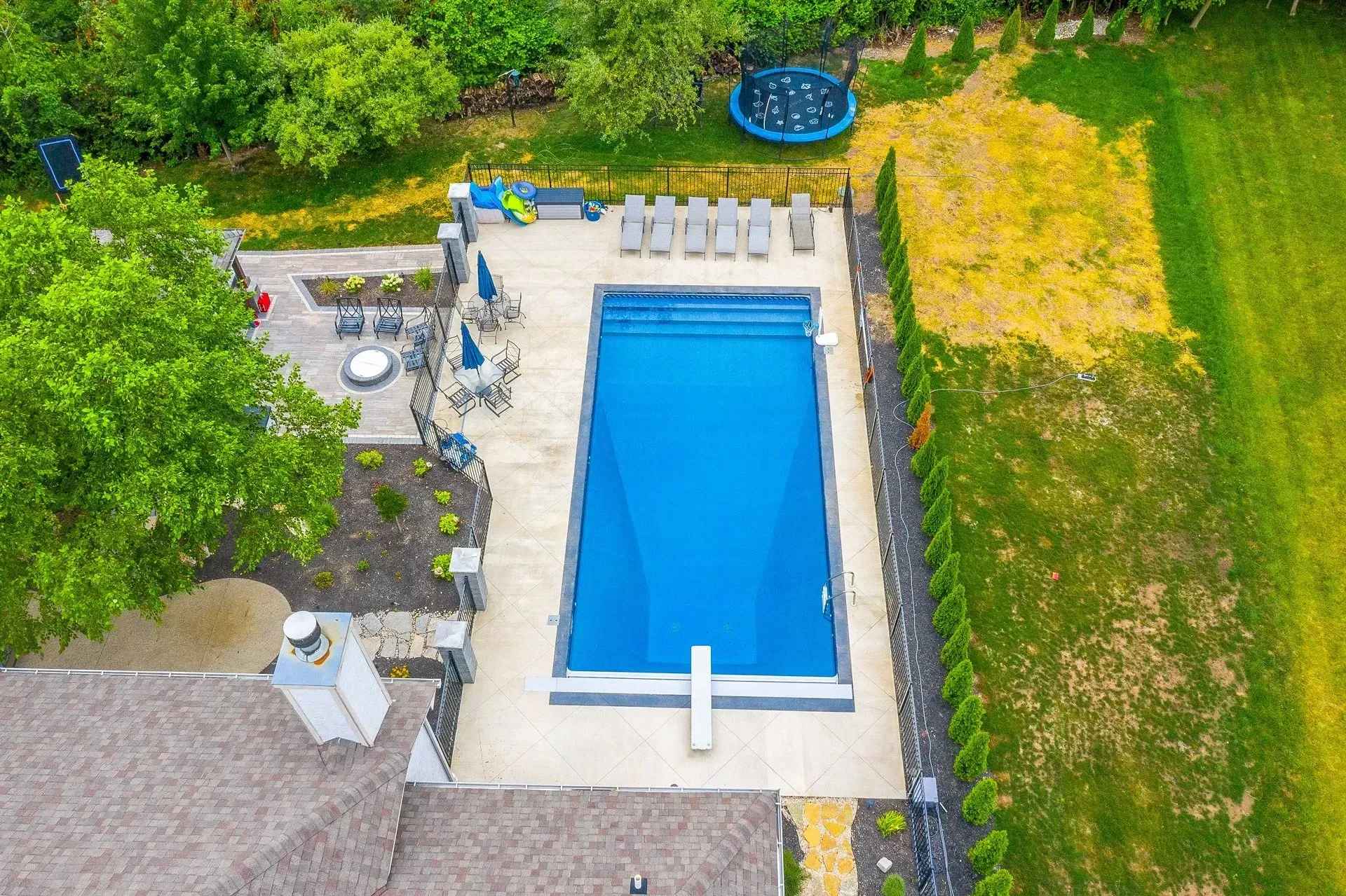 Aerial view of a rectangular in-ground swimming pool surrounded by patio furniture and a yard, a trampoline is in the background.