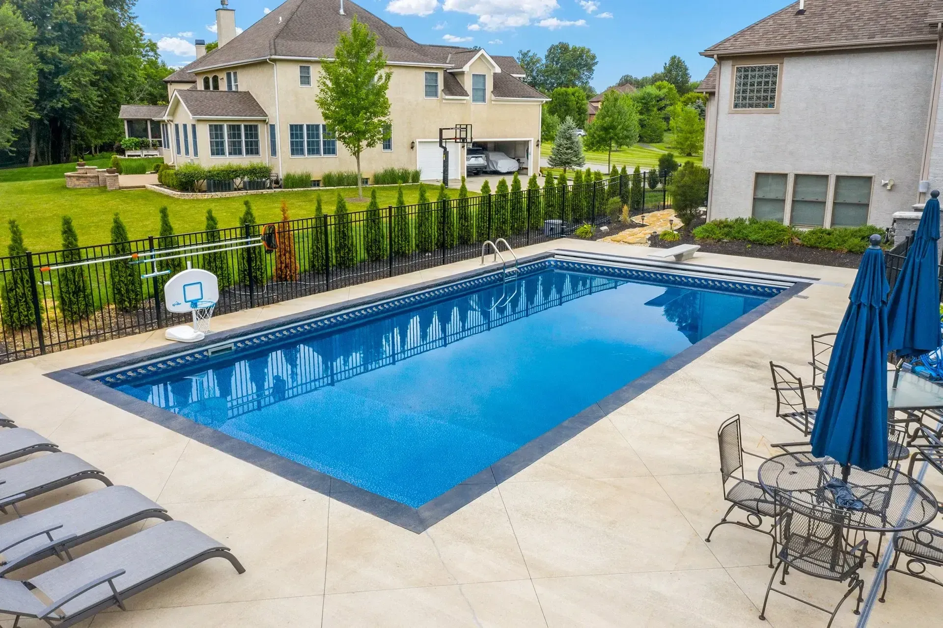 Backyard with rectangular pool, lounge chairs, tables, and a basketball hoop. The houses are in the background, and trees border the lawn.